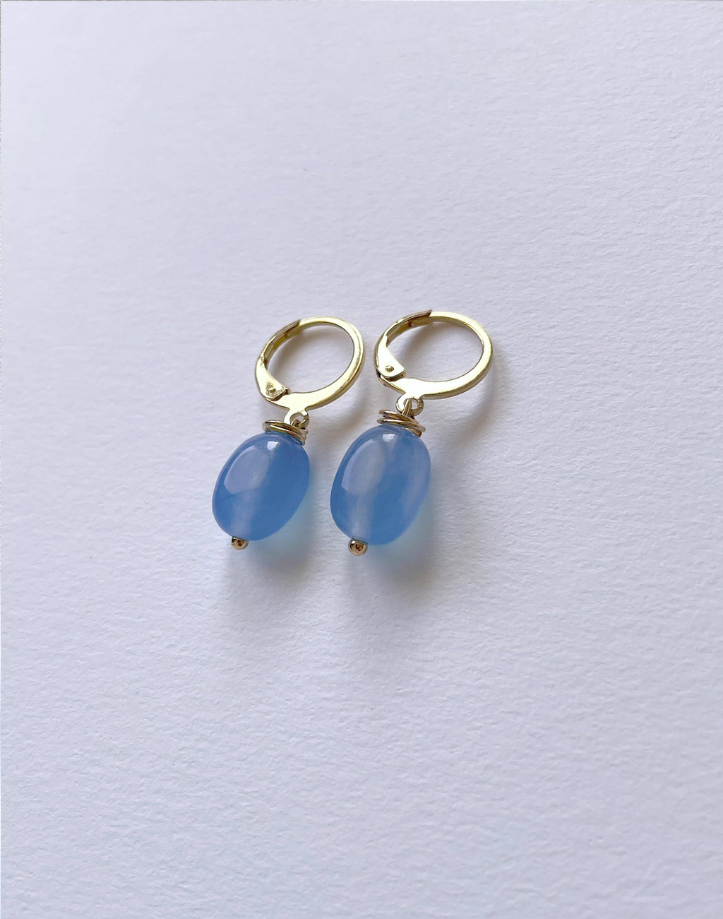 A pair of gold hoop earrings with smooth, oval light blue bead drops, displayed on a clean, light gray textured surface.