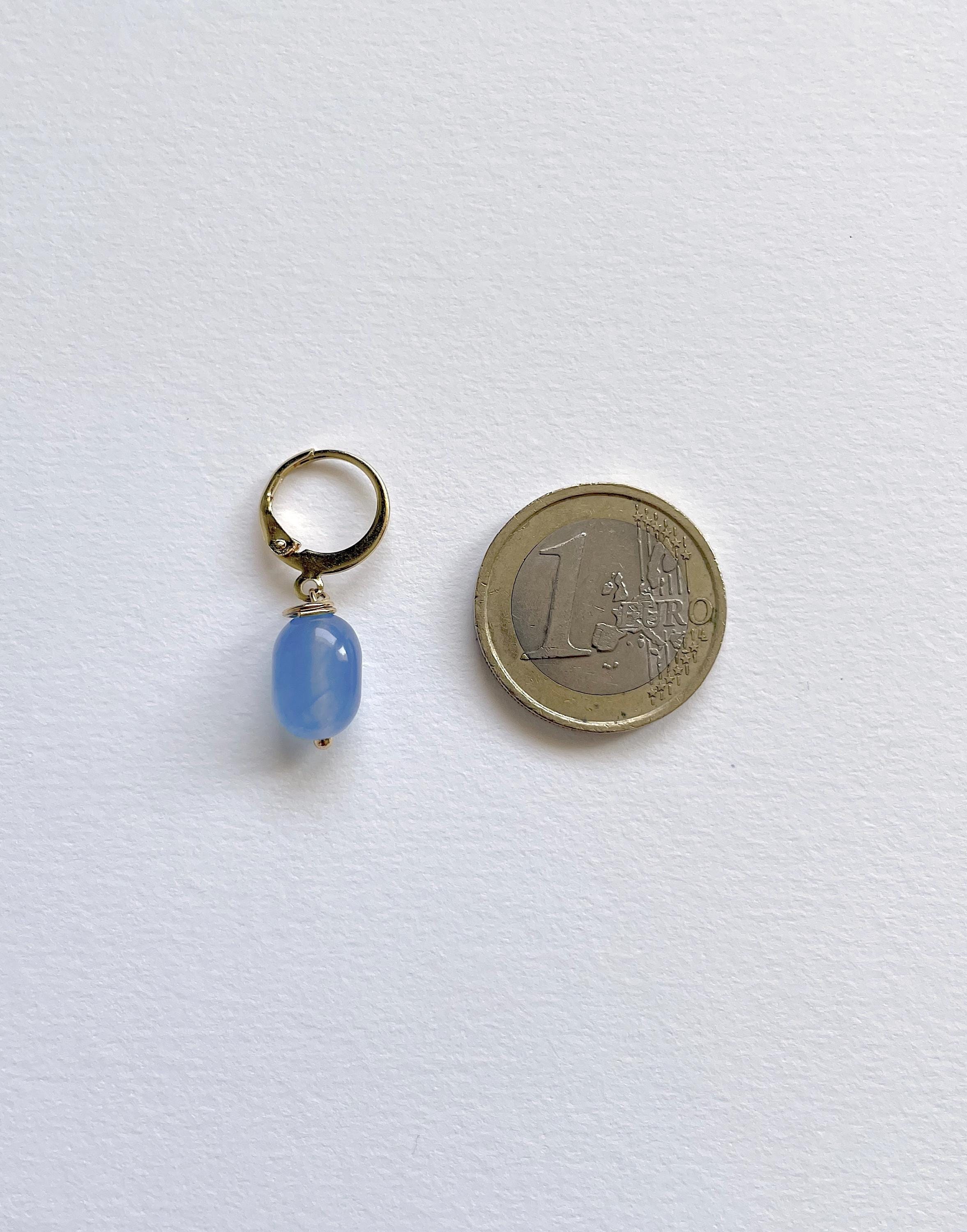 A small gold-tone hoop earring with a dangling light blue oval bead, shown next to a 1 Euro coin for scale on a white textured background.