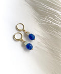 A pair of gold hoop earrings with small sage green amazonite beads and round blue jade beads dangling below, photographed on a white surface with soft white feather-like decor.
