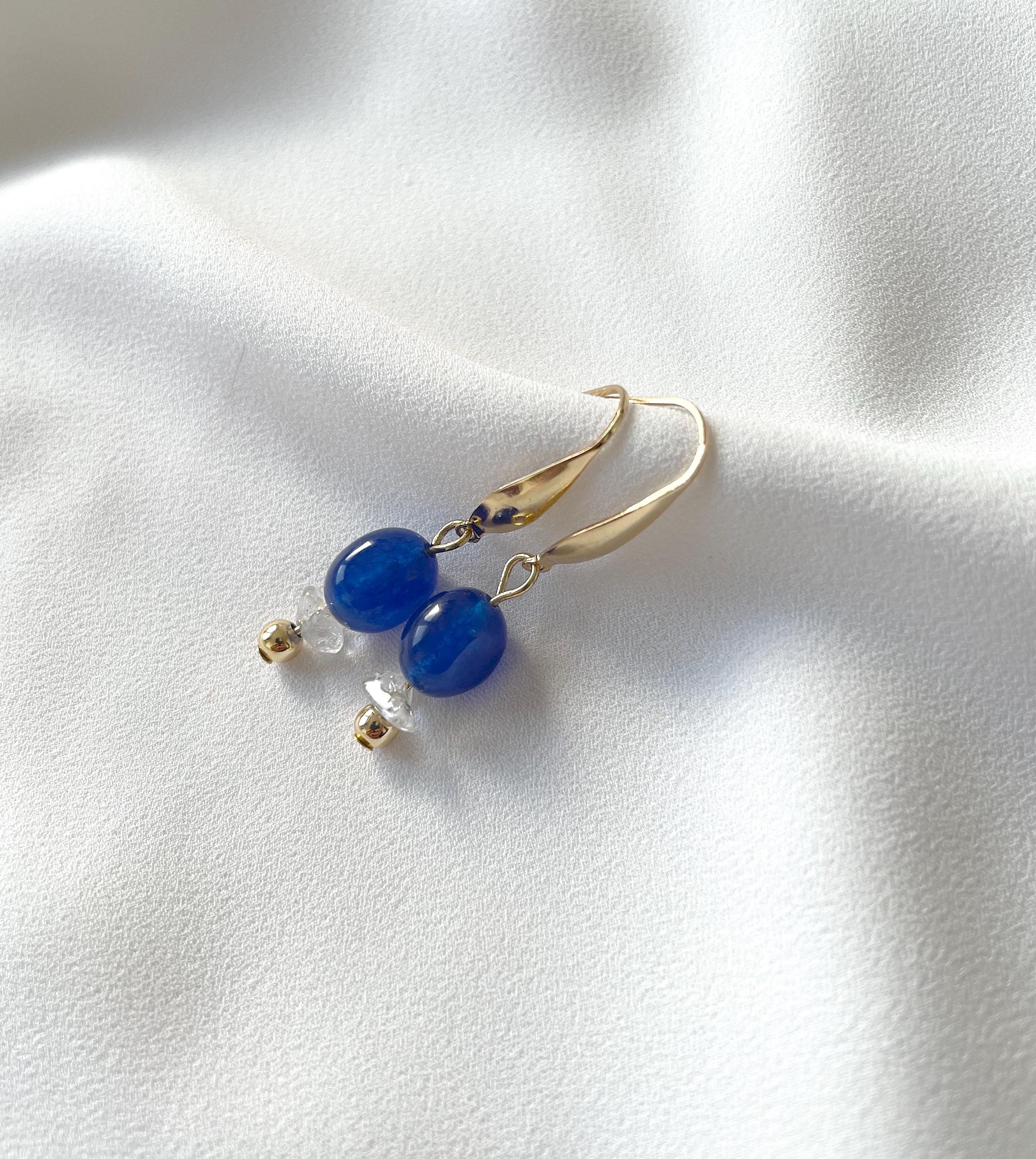 A pair of gold drop earrings with smooth blue oval beads and small clear crystal accents, displayed on a soft white fabric background.