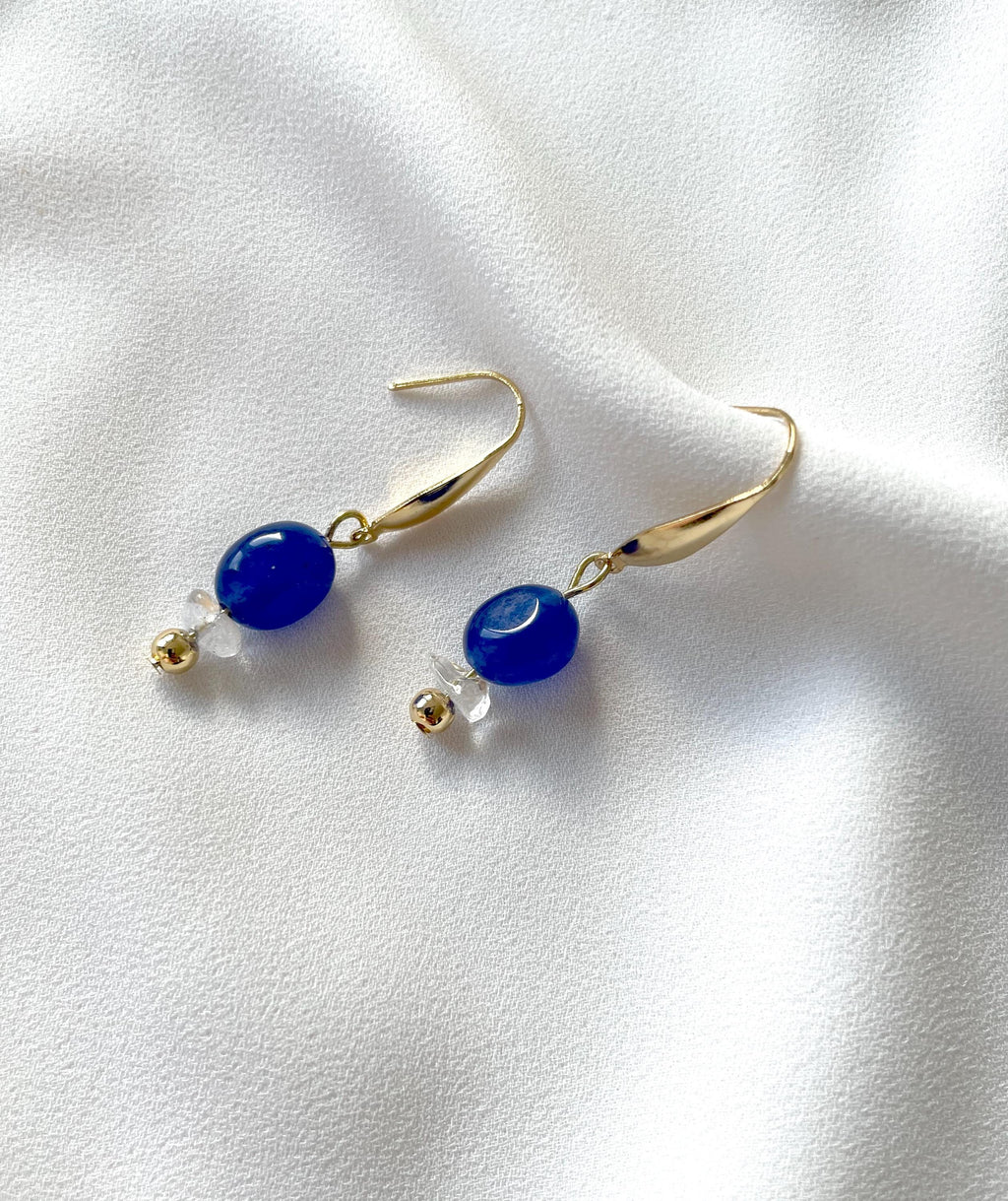 A pair of gold plated dangle and drop earrings with smooth blue oval jade beads and small clear quartz crystal accents, displayed on a soft white fabric background.