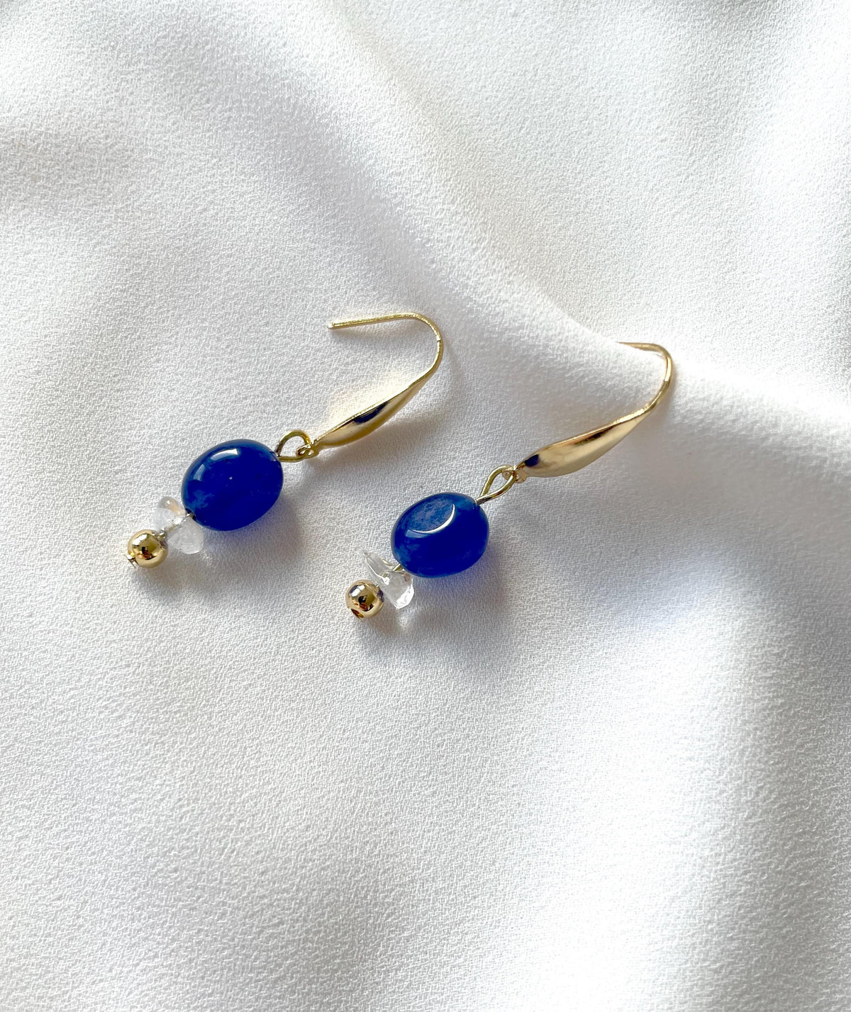 A pair of gold plated dangle and drop earrings with smooth blue oval jade beads and small clear quartz crystal accents, displayed on a soft white fabric background.