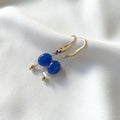 A pair of gold plated drop earrings with polished navy blue natural jade beads and small clear quartz crystal chip bead accents, displayed on a soft white fabric background.