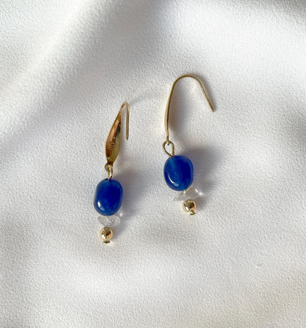A pair of gold plated dangle earrings with polished navy blue jade beads and small clear natural quartz chip bead accents, displayed on a textured white fabric background.