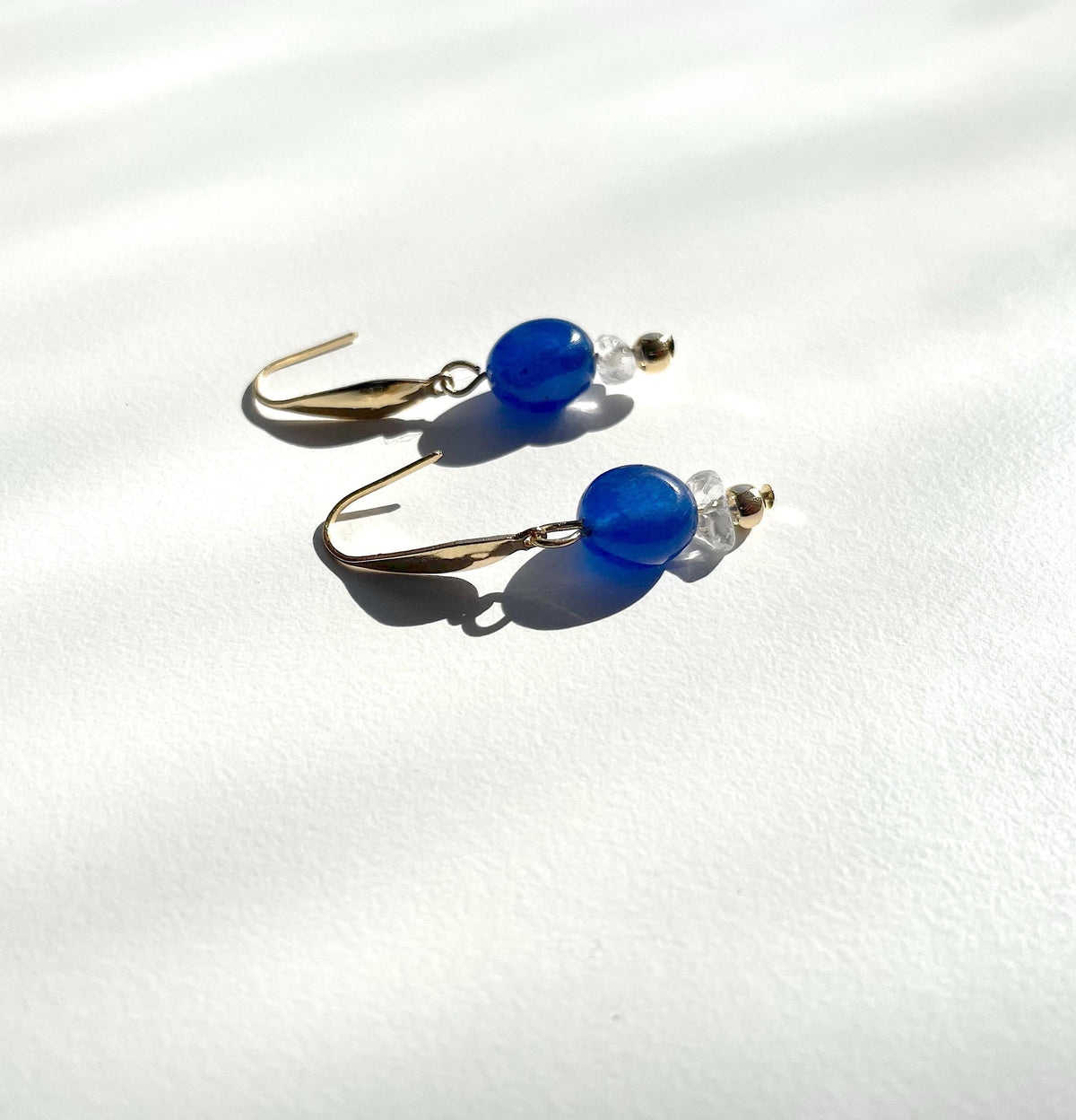 A pair of gold-tone gold plated dangle earrings with smooth, round blue jade beads and small clear crystal quartz chip bead accents, displayed on a textured white surface with soft shadows.