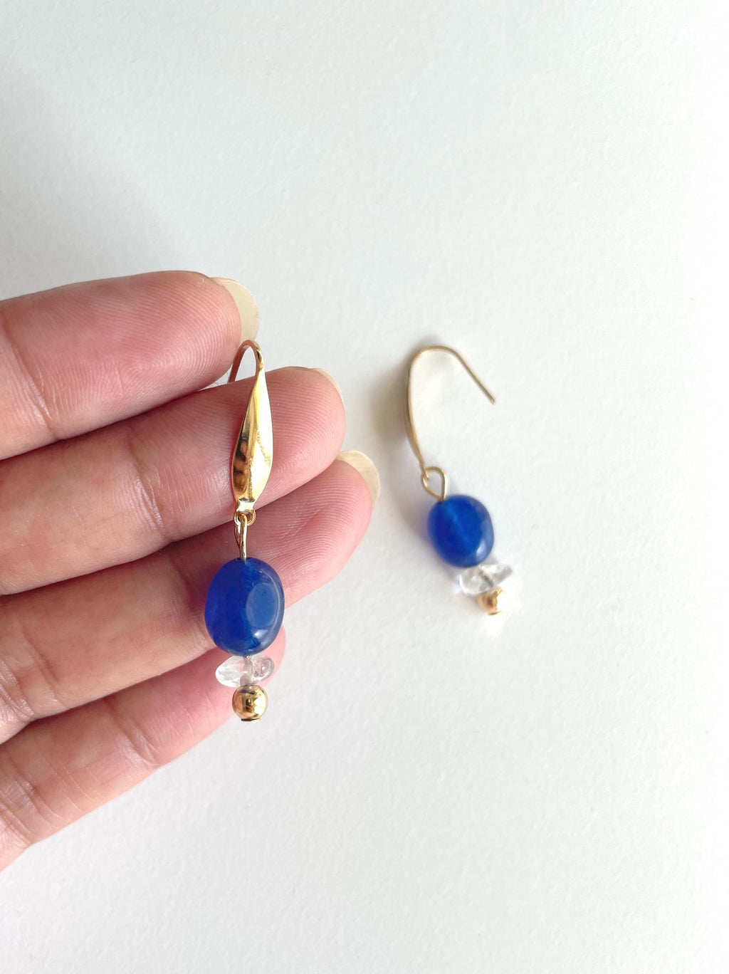 A pair of gold plated drop earrings with blue jade beads and small clear real quartz crystal chip bead accents, shown with one earring held by a hand against a plain white background.