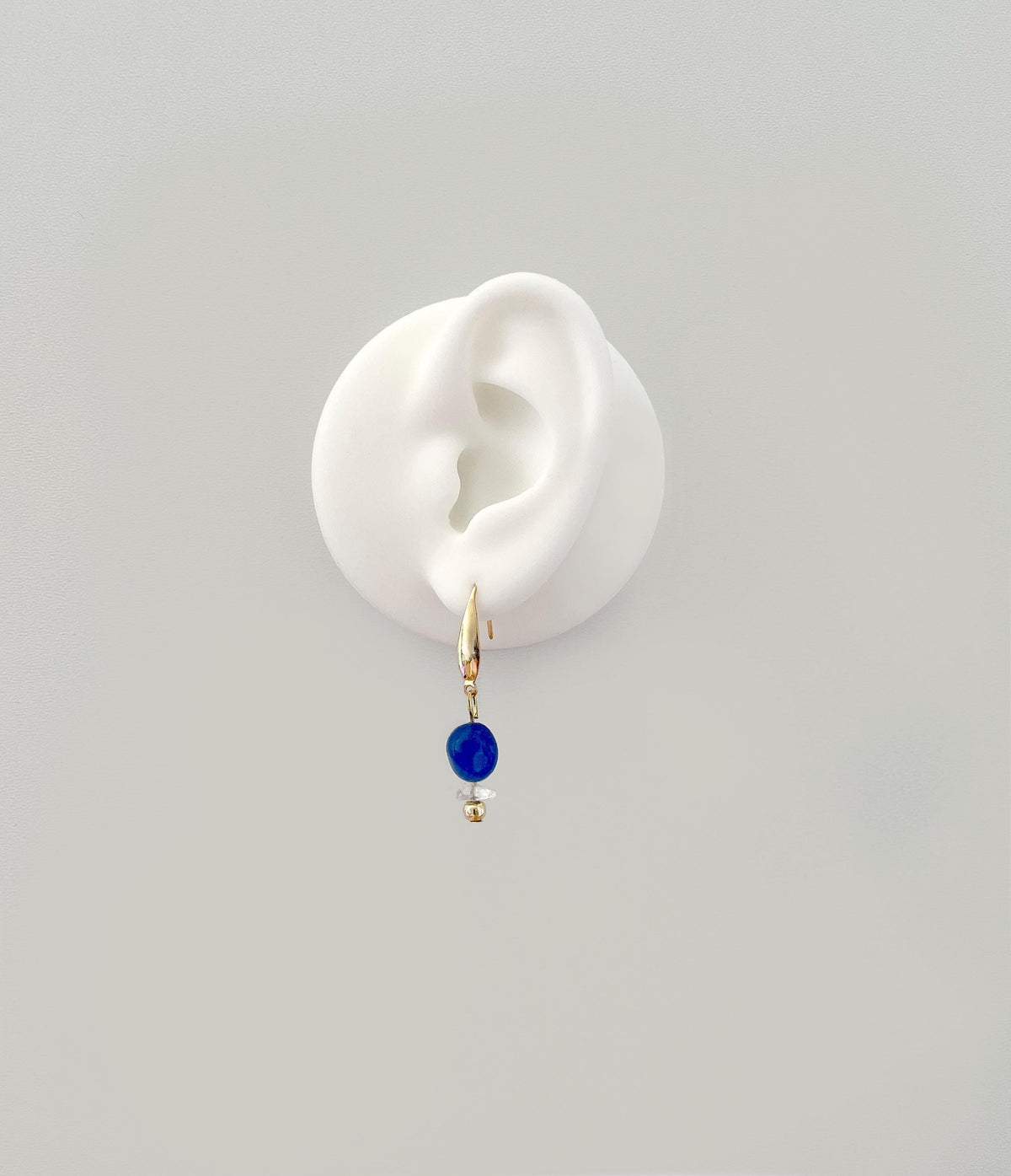 A single gold plated hoop earring with a small dark blue jade bead and tiny gold spacer, displayed on a white ear-shaped stand against a light gray background.