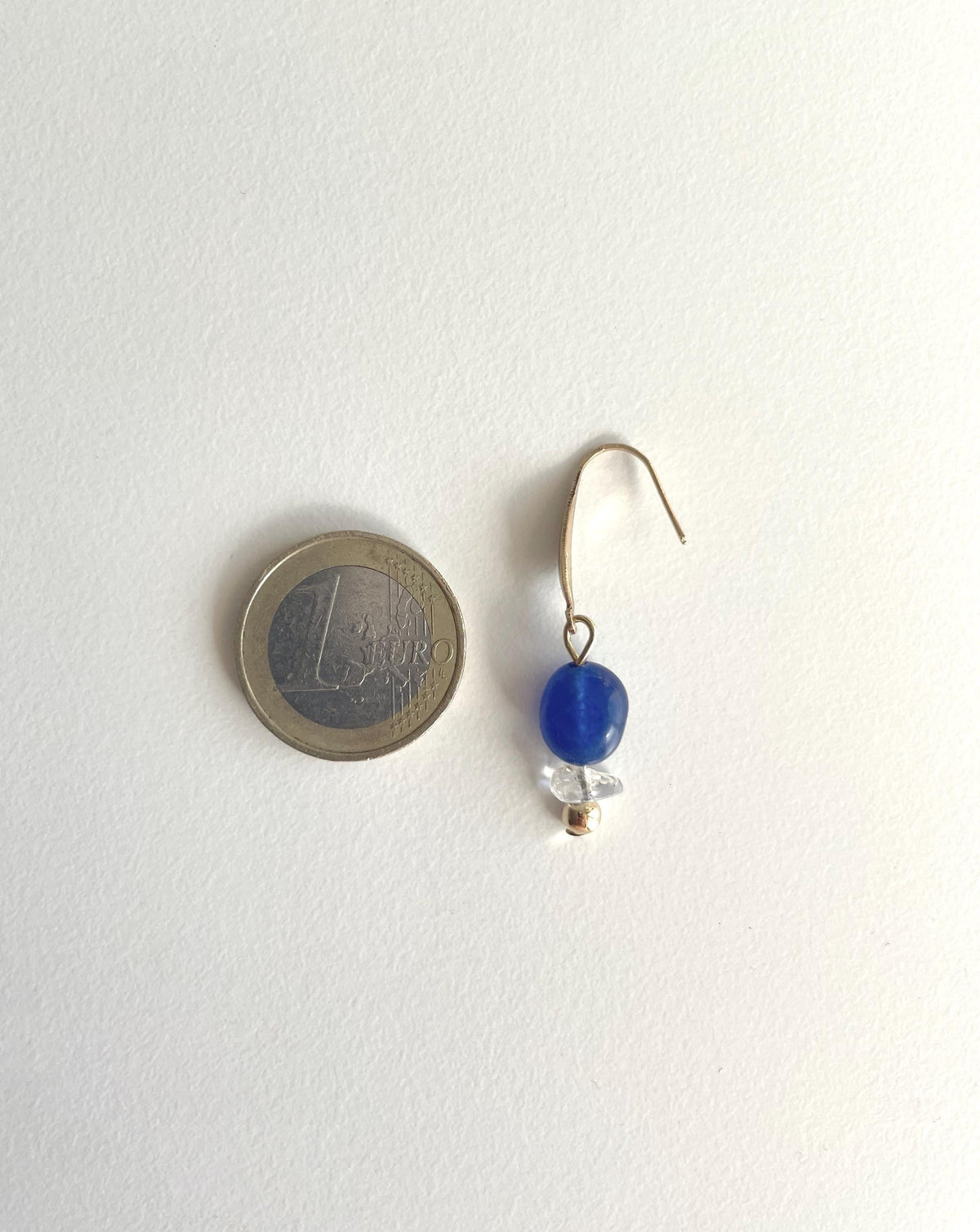A single small drop earring with a gold-tone hook, a round blue jade bead, and a small clear faceted quartz chip bead, shown next to a 1 Euro coin for scale on a plain white background.