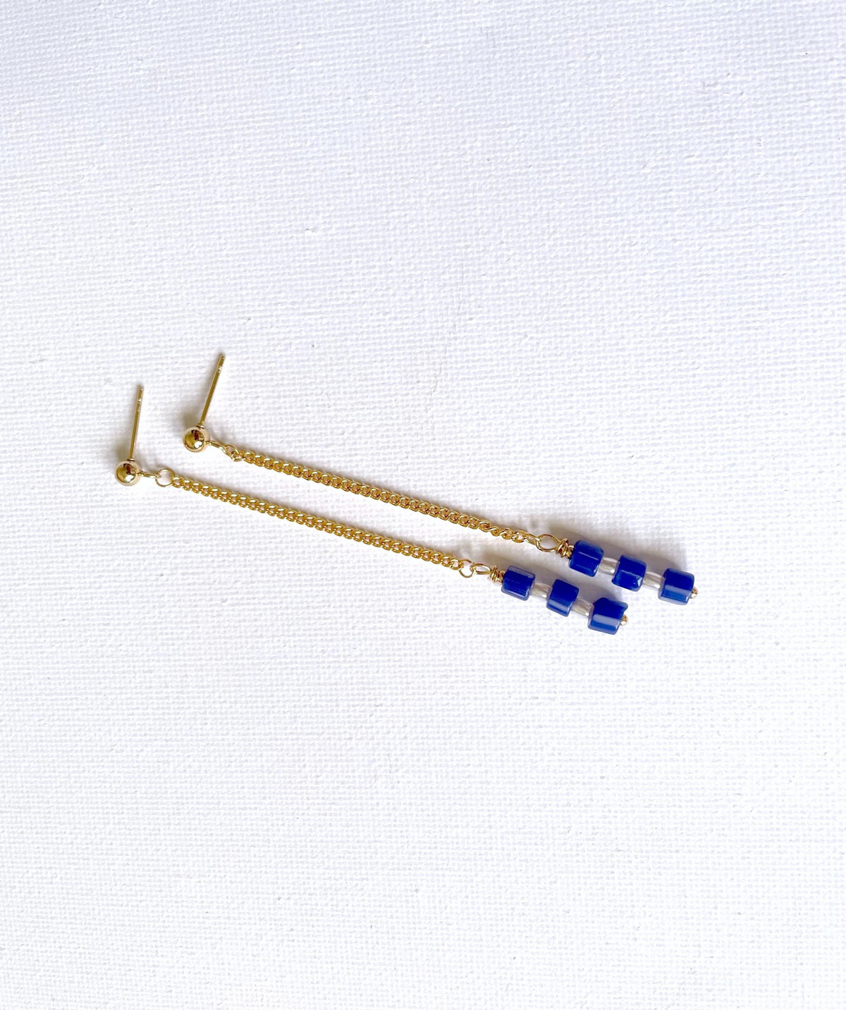 A pair of gold-toned dangle threader earrings with small blue beads and real white pearls, displayed on a textured white background.