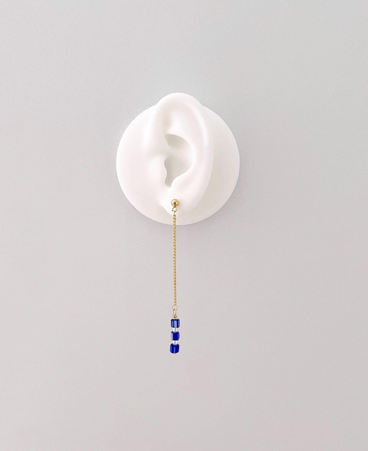 A single dangling earring displayed on a white ear-shaped stand against a light gray background. The earring has a gold chain and small blue beads with white pearls at the end.
