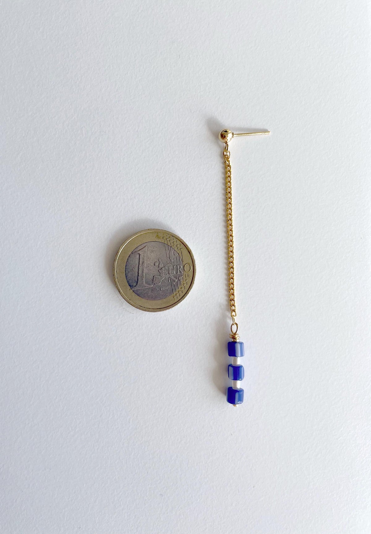 A single gold-toned dangle earring with three small blue beads at the end, shown next to a 1 euro coin for scale on a plain white background.
