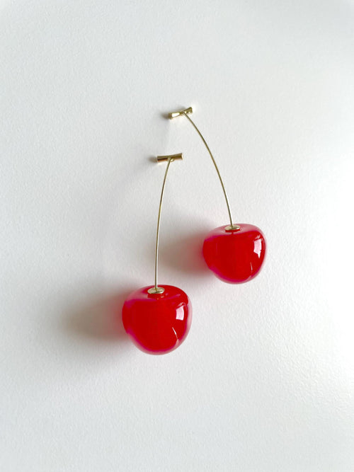 A pair of dangling earrings shaped like glossy red cherries with thin gold stems and small gold posts, shown against a plain white wall.