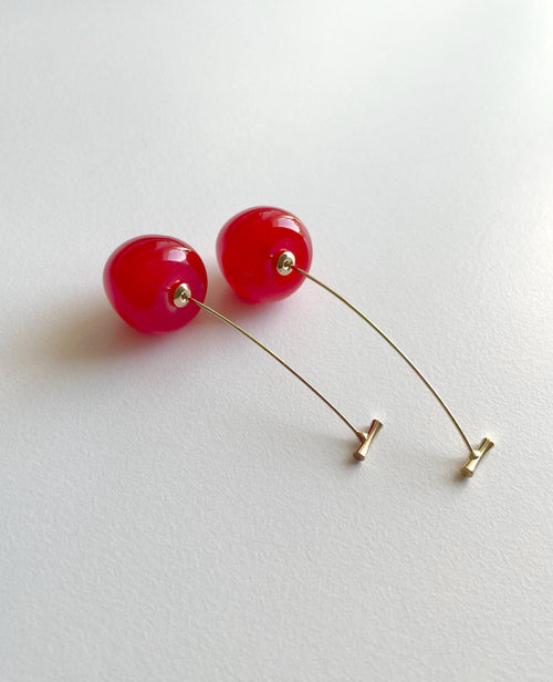A pair of long, minimalist drop earrings featuring glossy, spherical bright red beads suspended from thin gold wire posts with small gold end caps, displayed on a clean white surface.
