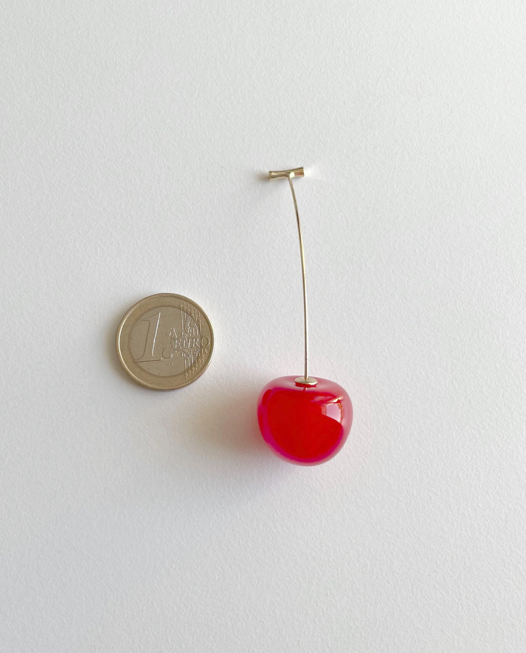 A miniature red cherry earring with a gold-tone post, shown next to a 1 Euro coin for scale on a plain white background.