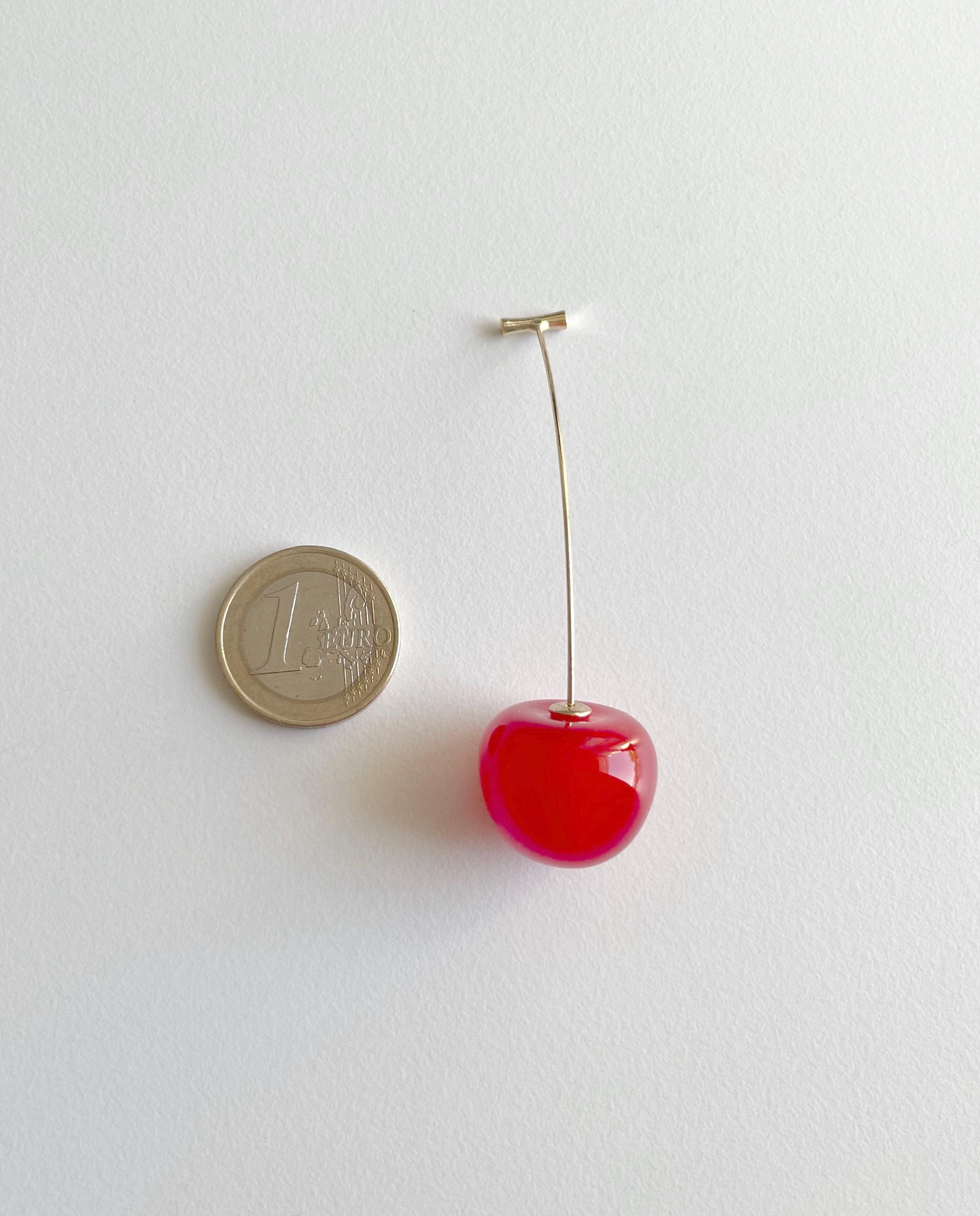 A miniature red cherry earring with a gold-tone post, shown next to a 1 Euro coin for scale on a plain white background.