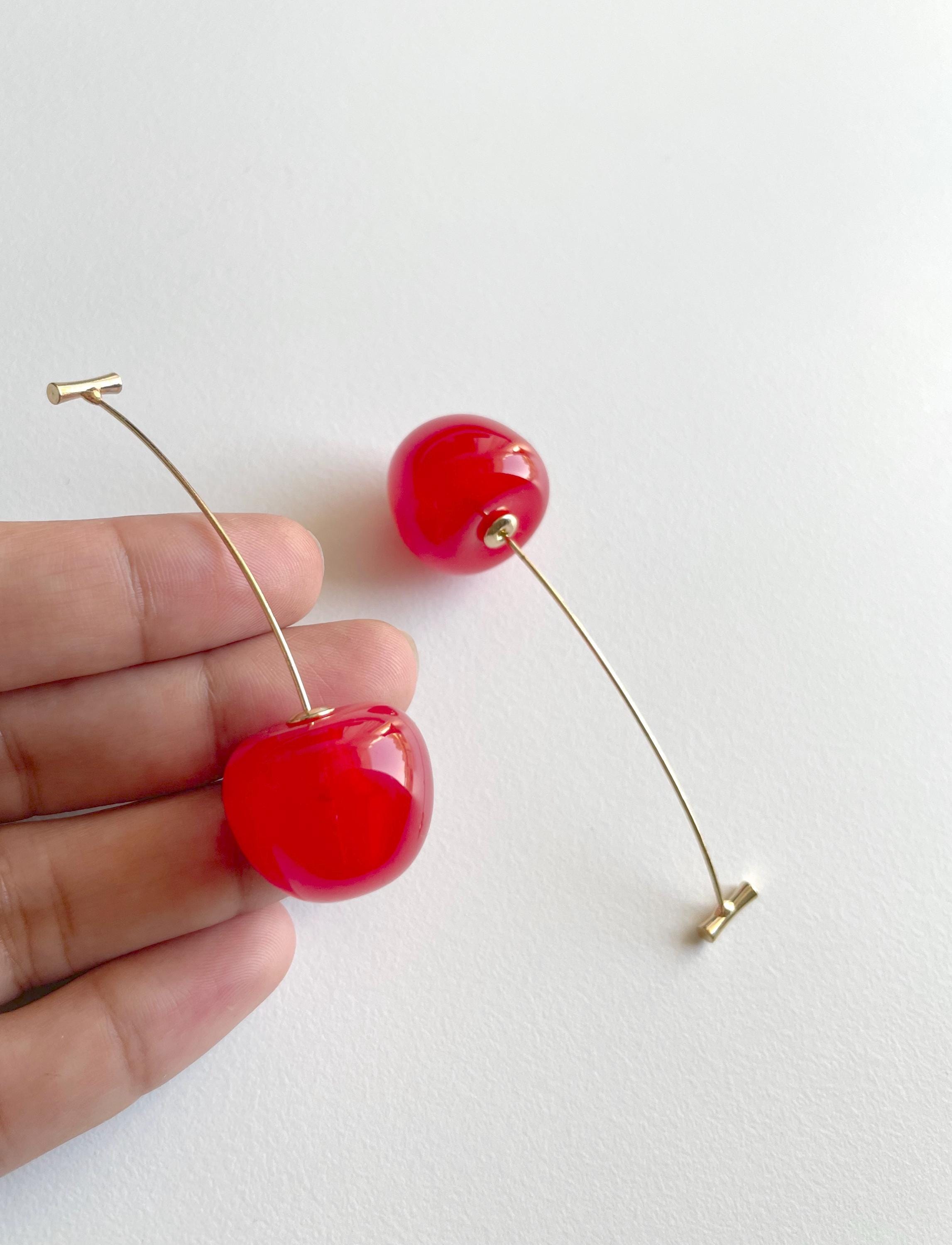 A pair of cherry-shaped drop earrings with glossy red resin cherries and gold-tone wire stems, shown with a person's fingers holding one earring against a plain white background.