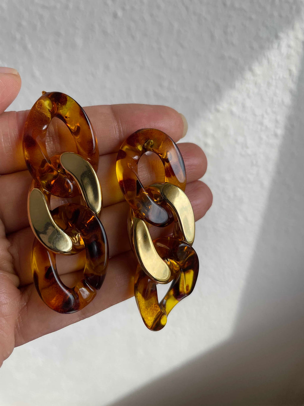 A pair of chunky interlocking chain-link earrings in a translucent amber tortoiseshell pattern with shiny gold-toned accents, held in a hand against a white textured wall.