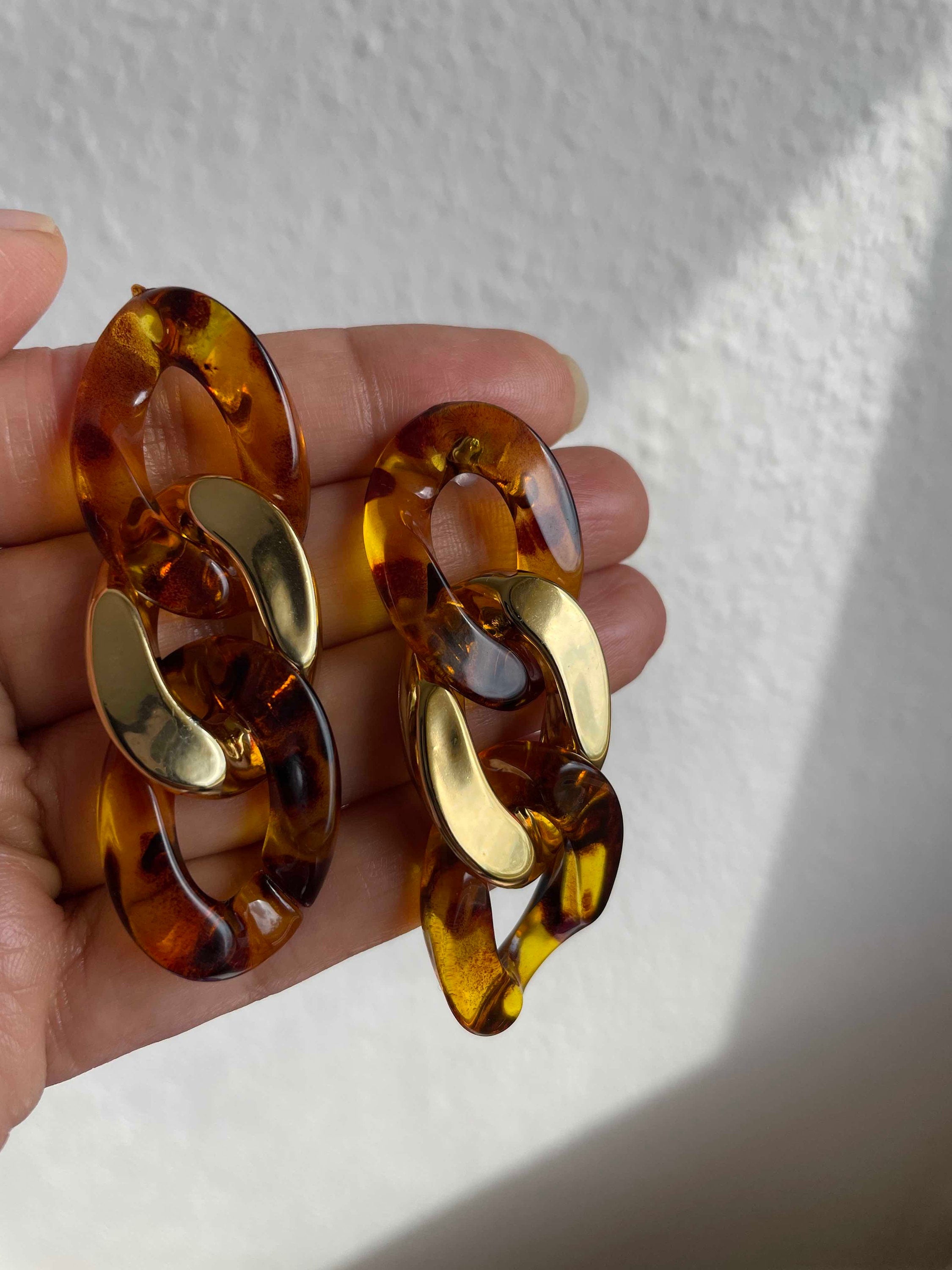 A pair of chunky interlocking chain-link earrings in a translucent amber tortoiseshell pattern with shiny gold-toned accents, held in a hand against a white textured wall.