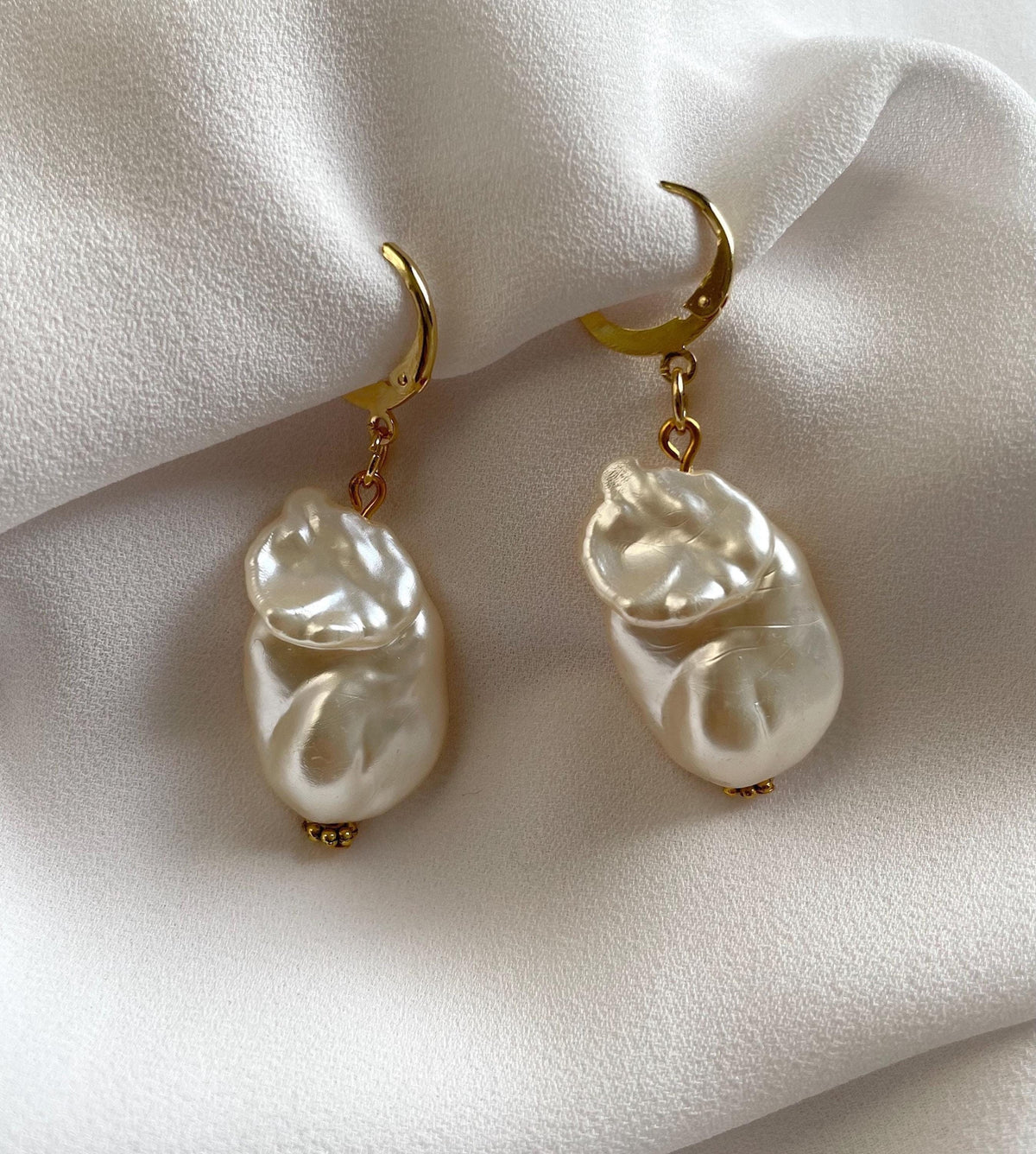 A pair of gold plated drop earrings with irregular white baroque pearls, displayed on a soft white fabric background.
