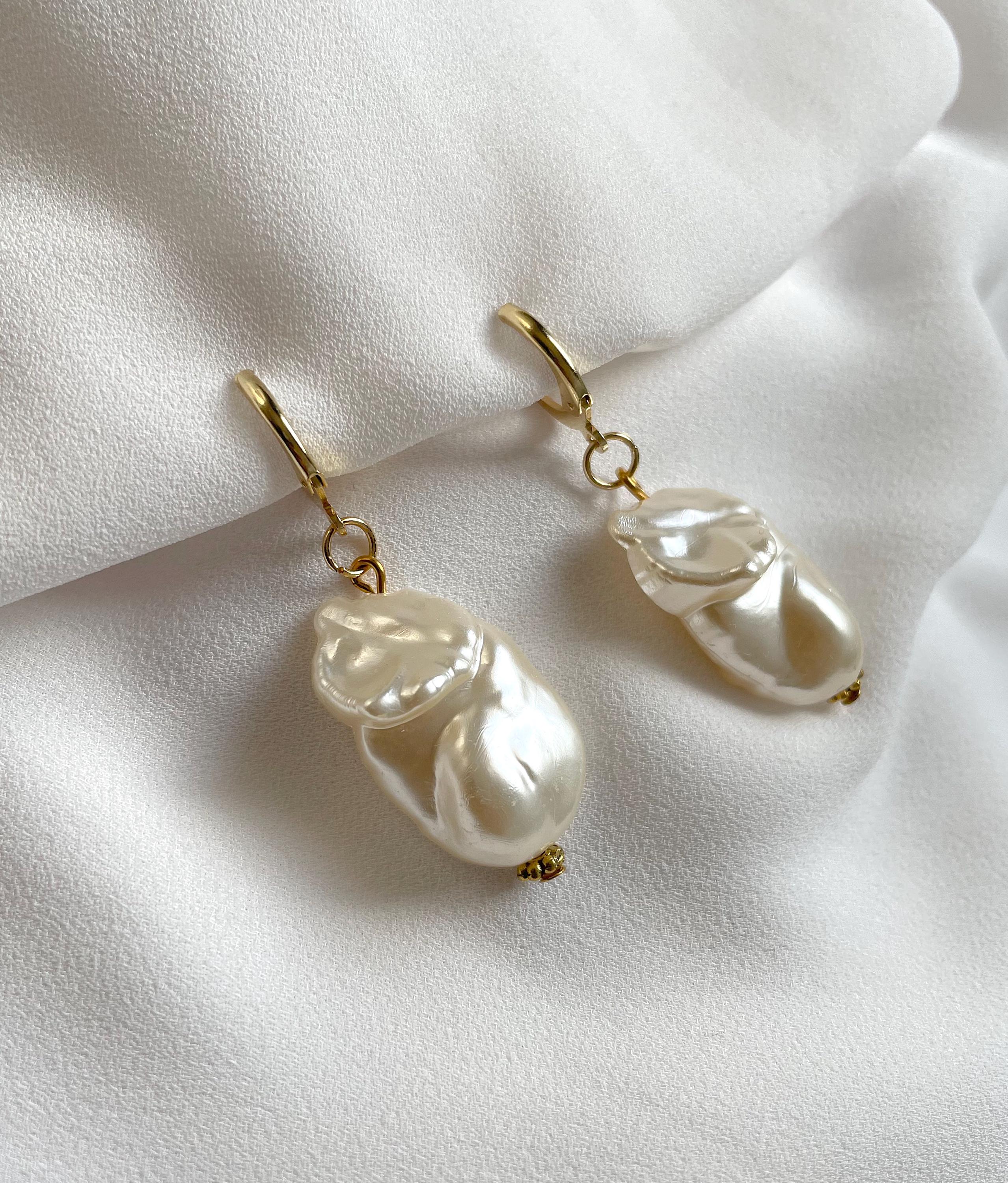 A pair of gold huggie hoop earrings with irregular white baroque pearls dangling from gold rings, displayed on a soft white fabric background.