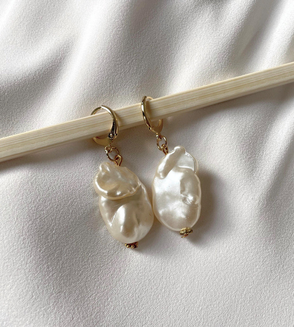 A pair of gold hoop earrings with irregular, lustrous white baroque pearls, displayed on a light wood chopstick against a textured white fabric background.
