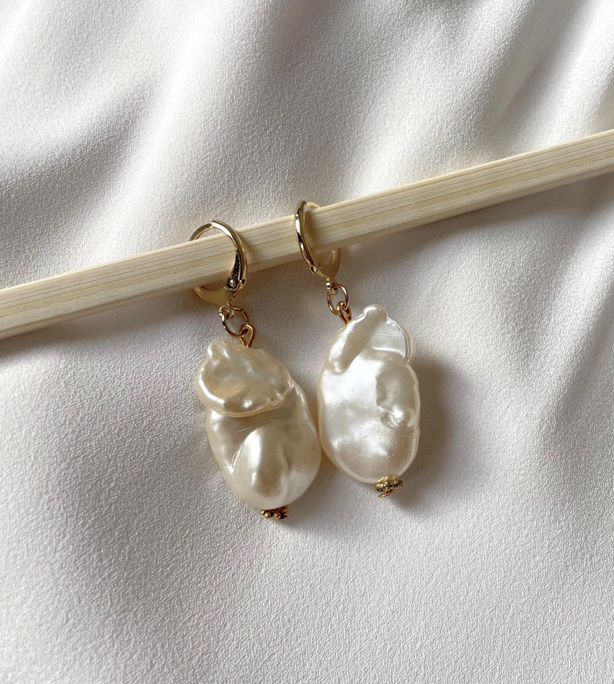 A pair of gold hoop earrings with irregular, lustrous white baroque pearls, displayed on a light wood chopstick against a textured white fabric background.