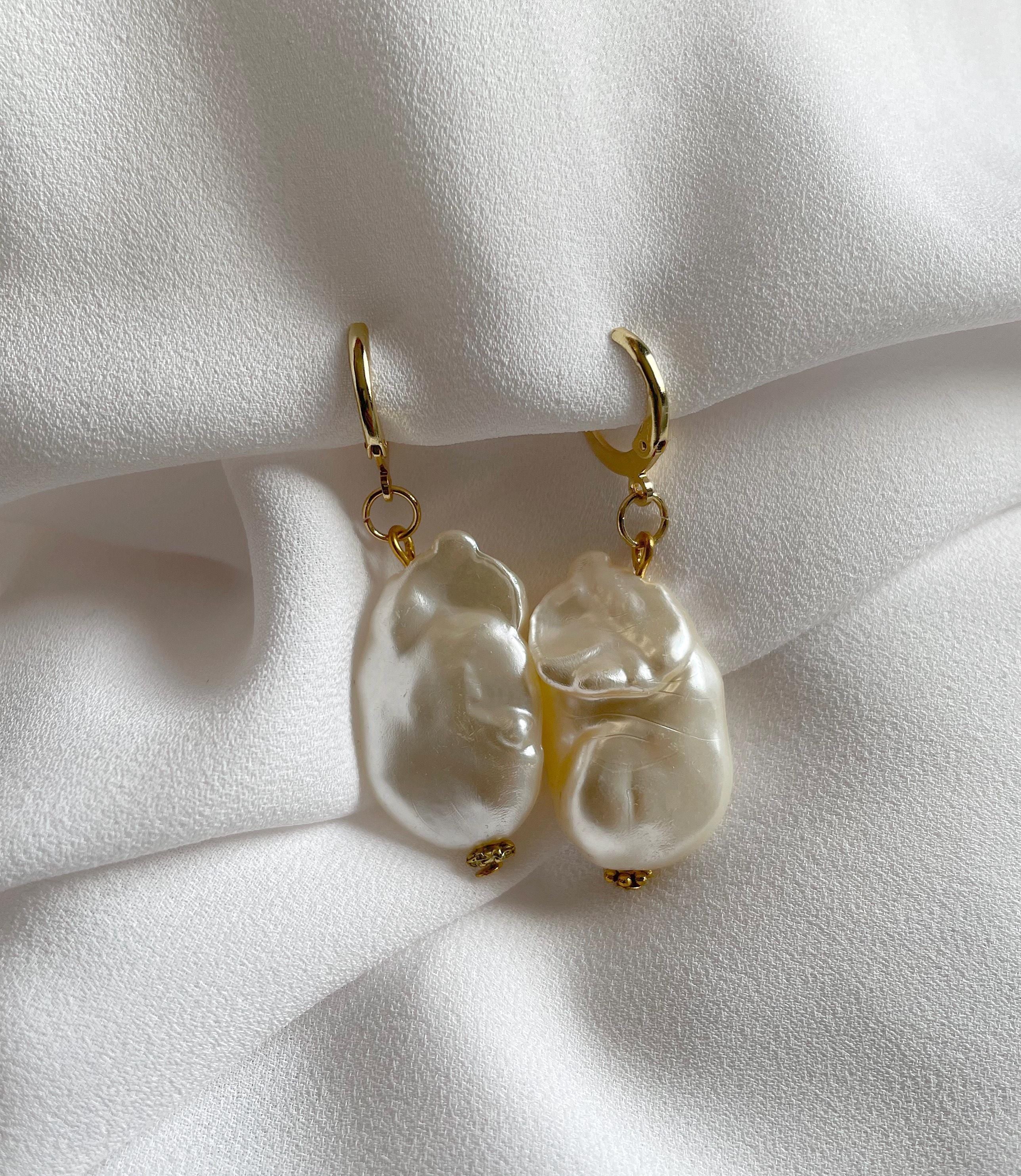 A pair of gold hoop earrings with large, irregular white baroque pearls, displayed on a soft, draped white fabric background.