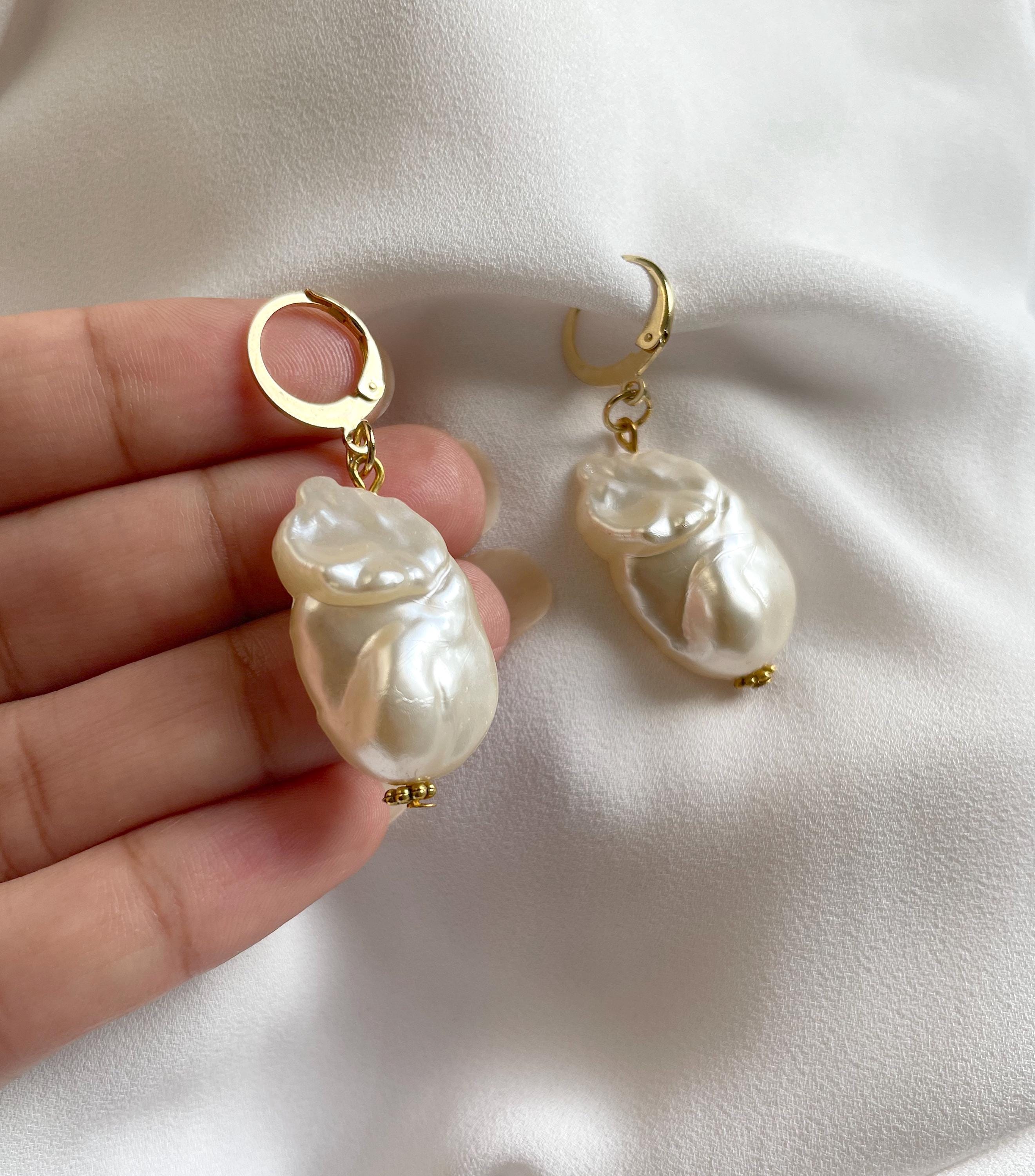 A pair of gold hoop earrings with large, irregular white baroque pearls hanging from gold rings, held in a person's hand against a white fabric background.