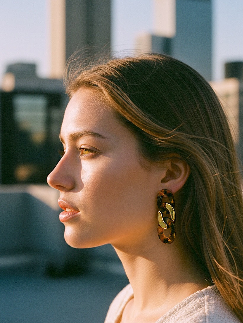 A woman in profile wearing large, chunky geometric drop earrings with a brown and black tortoiseshell pattern and gold-toned metal accents, set against a blurred cityscape background.