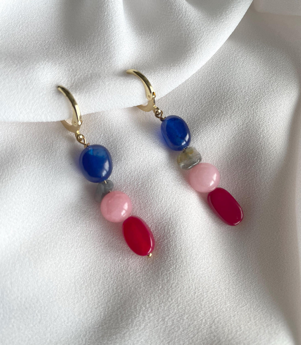 A pair of gold-tone dangle earrings with colorful gemstone beads, displayed on a soft white fabric background. The earrings feature a blue bead, a light pink bead, a dark red bead, and a small clear stone spacer.