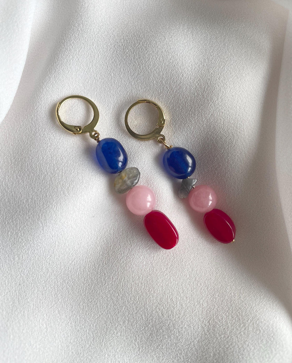 A pair of drop earrings with gold-tone hoops and colorful gemstone beads, including blue, pink, red, and gray, displayed on a white textured fabric background.