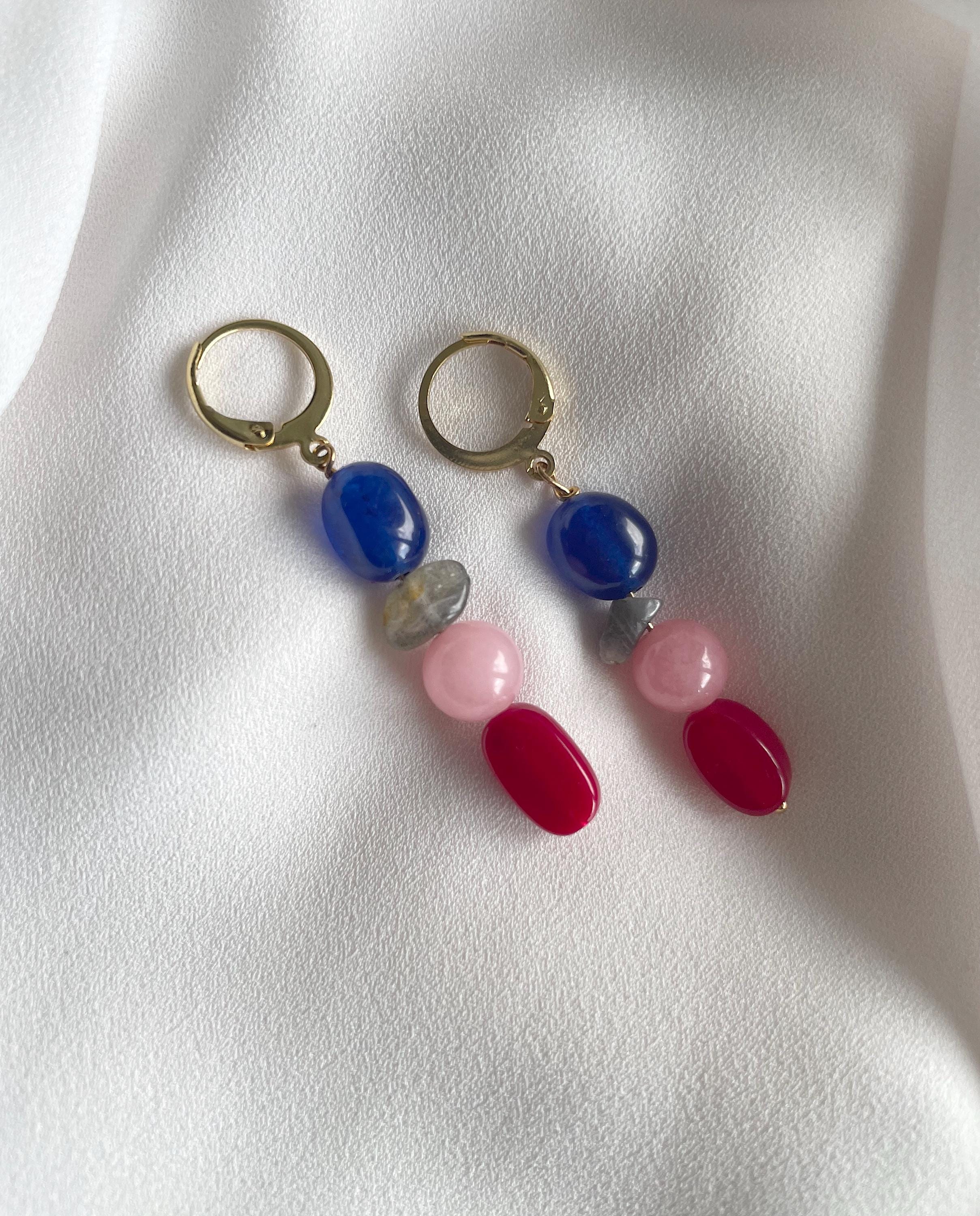 A pair of drop earrings with gold-tone hoops and colorful gemstone beads, including blue, pink, red, and gray, displayed on a white textured fabric background.