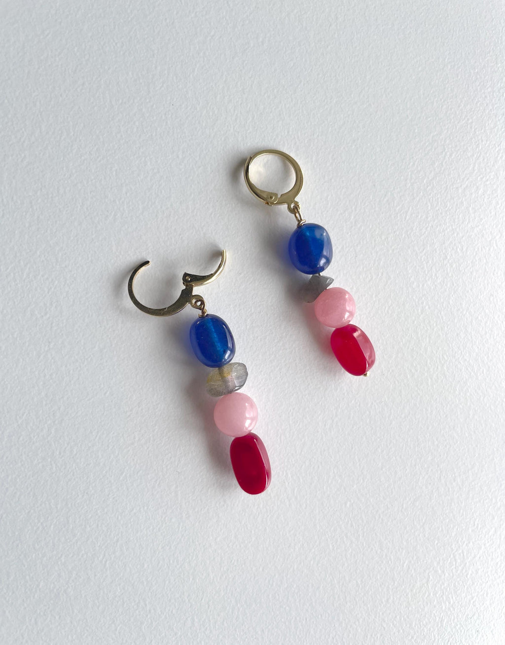 A pair of dangling earrings with antique gold-tone hoops and natural gemstone beads in blue, pink, red, and gray, displayed on a textured white surface.