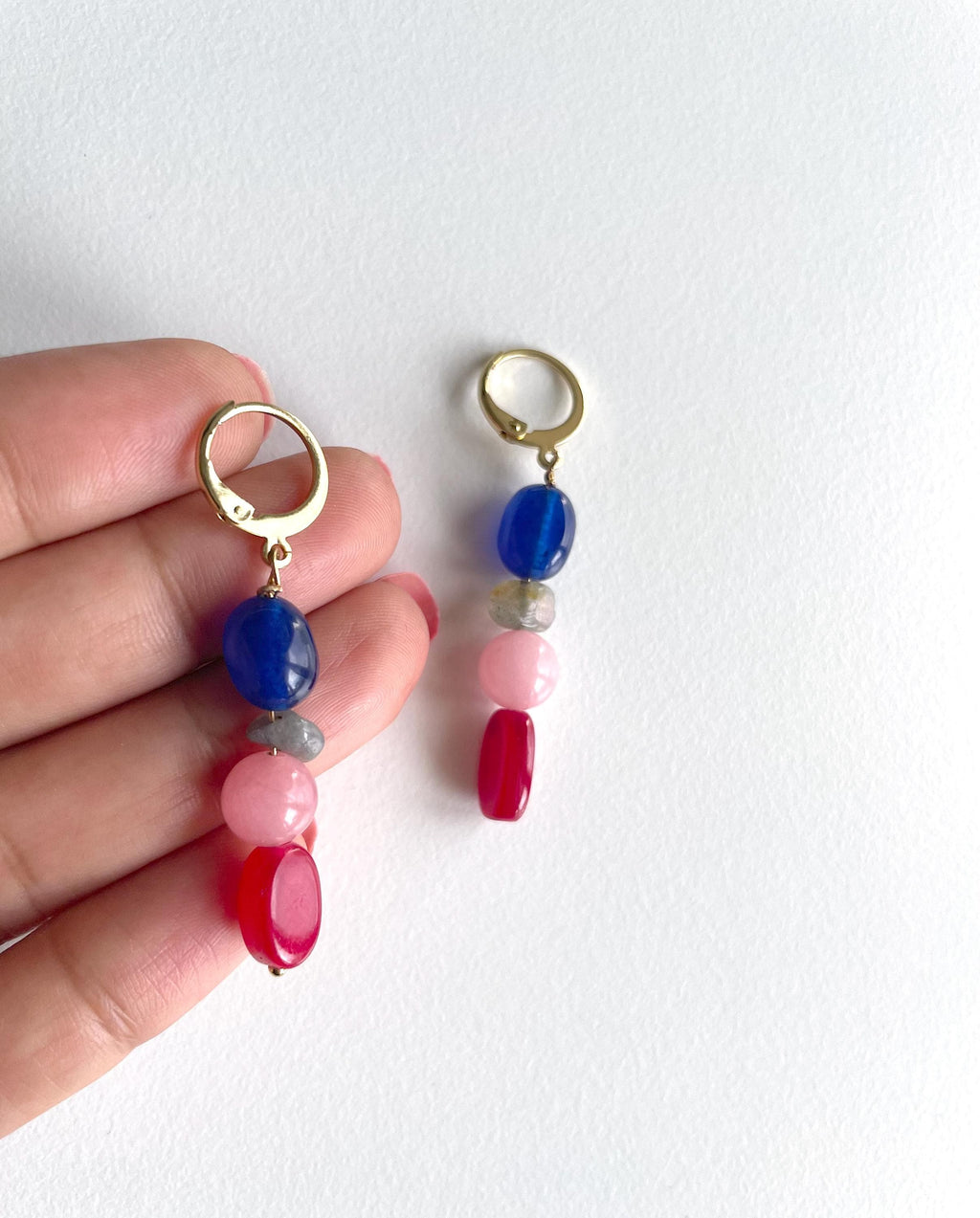 A pair of gold hoop drop earrings with colorful gemstone beads, held by a hand against a plain white background. The earrings feature a gold hoop, a dark blue bead, a light gray bead, a light pink bead, a dark red bead, and a large oval red bead at the bottom.