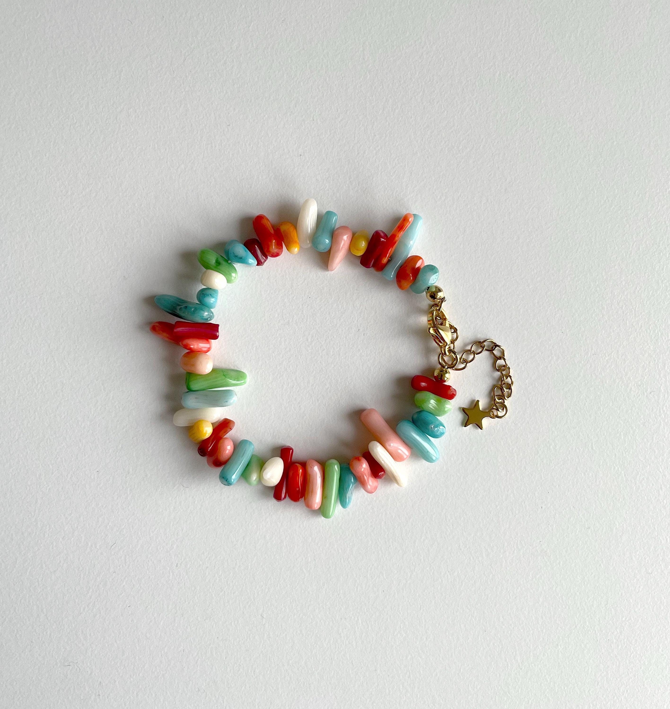 A multicolor beaded bracelet made of small cylindrical and irregular beads in red, blue, green, pink, yellow, and white, with a gold-tone chain and star charm, displayed on a light gray background.