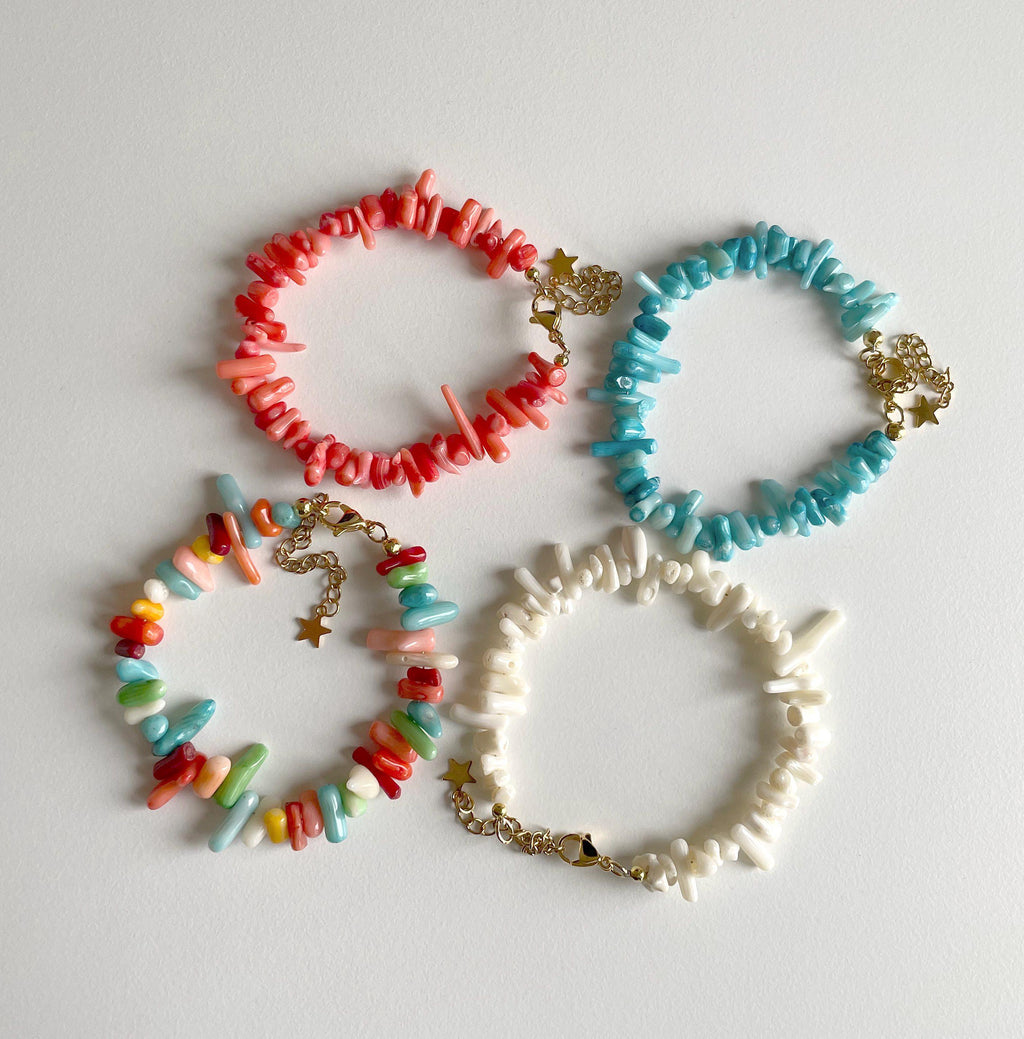 Four beaded stretch bracelets arranged on a light gray background. The set includes coral red, light blue, multicolor, and off-white bracelets, each decorated with small gold star charms.