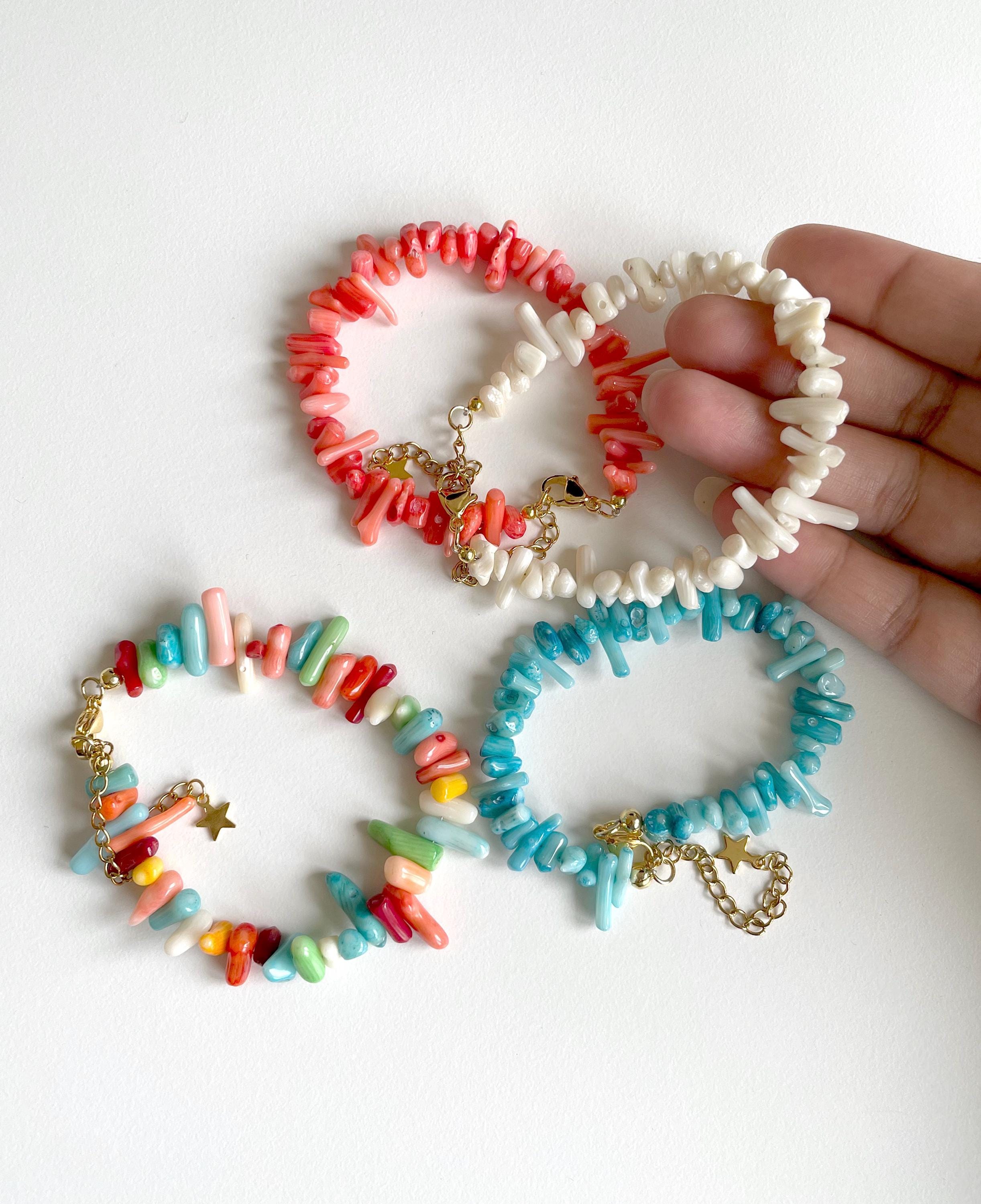 Four colorful beaded bracelets with gold charms are displayed on a white surface, with a hand holding two of the bracelets.