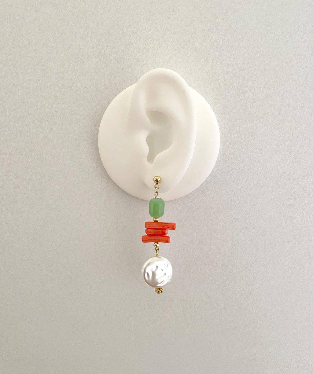 A single drop earring displayed on a white ear-shaped stand against a light gray background. The earring features a gold-tone post with a light green bead, stacked coral-red beads, and a white textured pearl dangle.