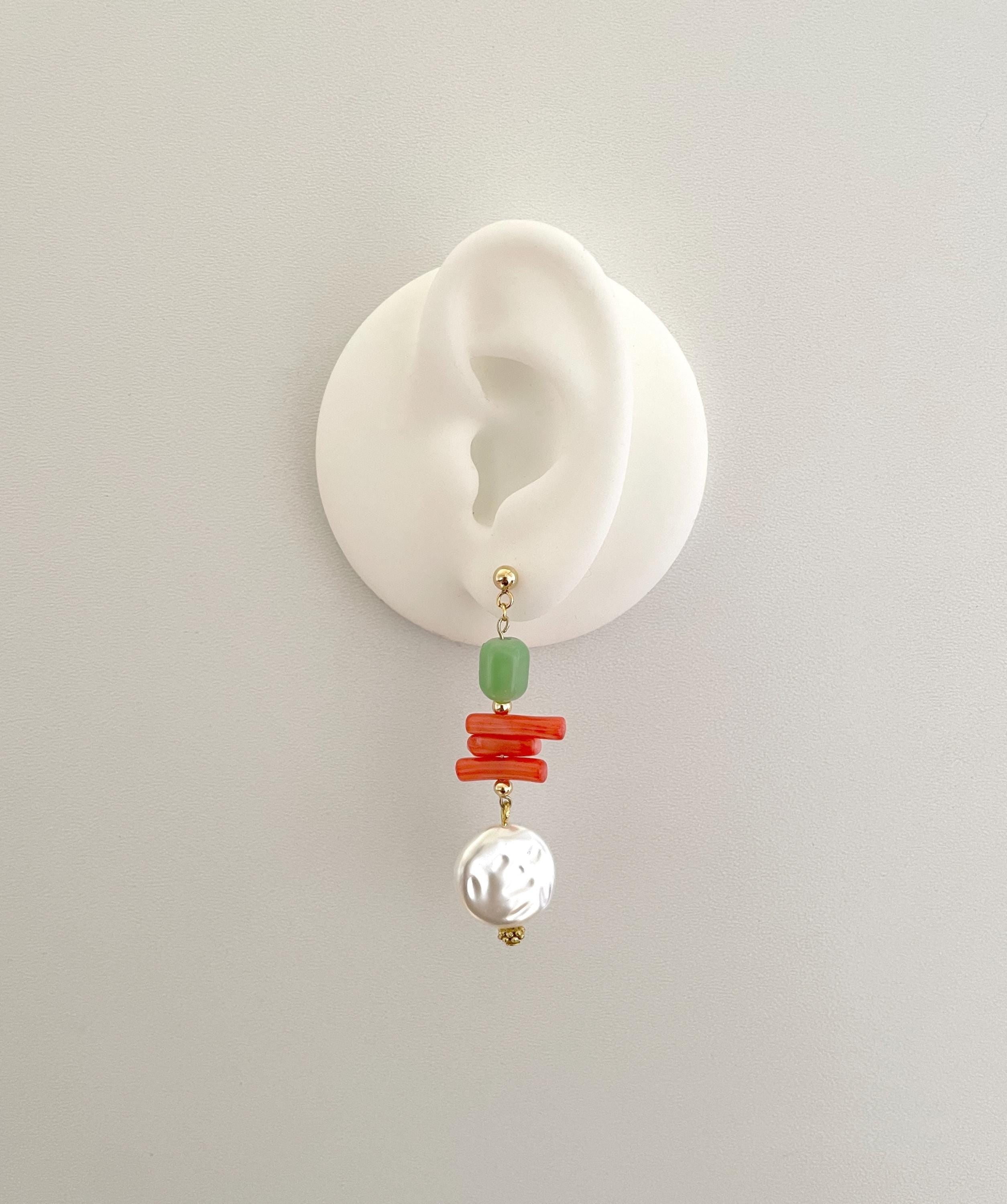 A single drop earring displayed on a white ear-shaped stand against a light gray background. The earring features a gold-tone post with a light green bead, stacked coral-red beads, and a white textured pearl dangle.