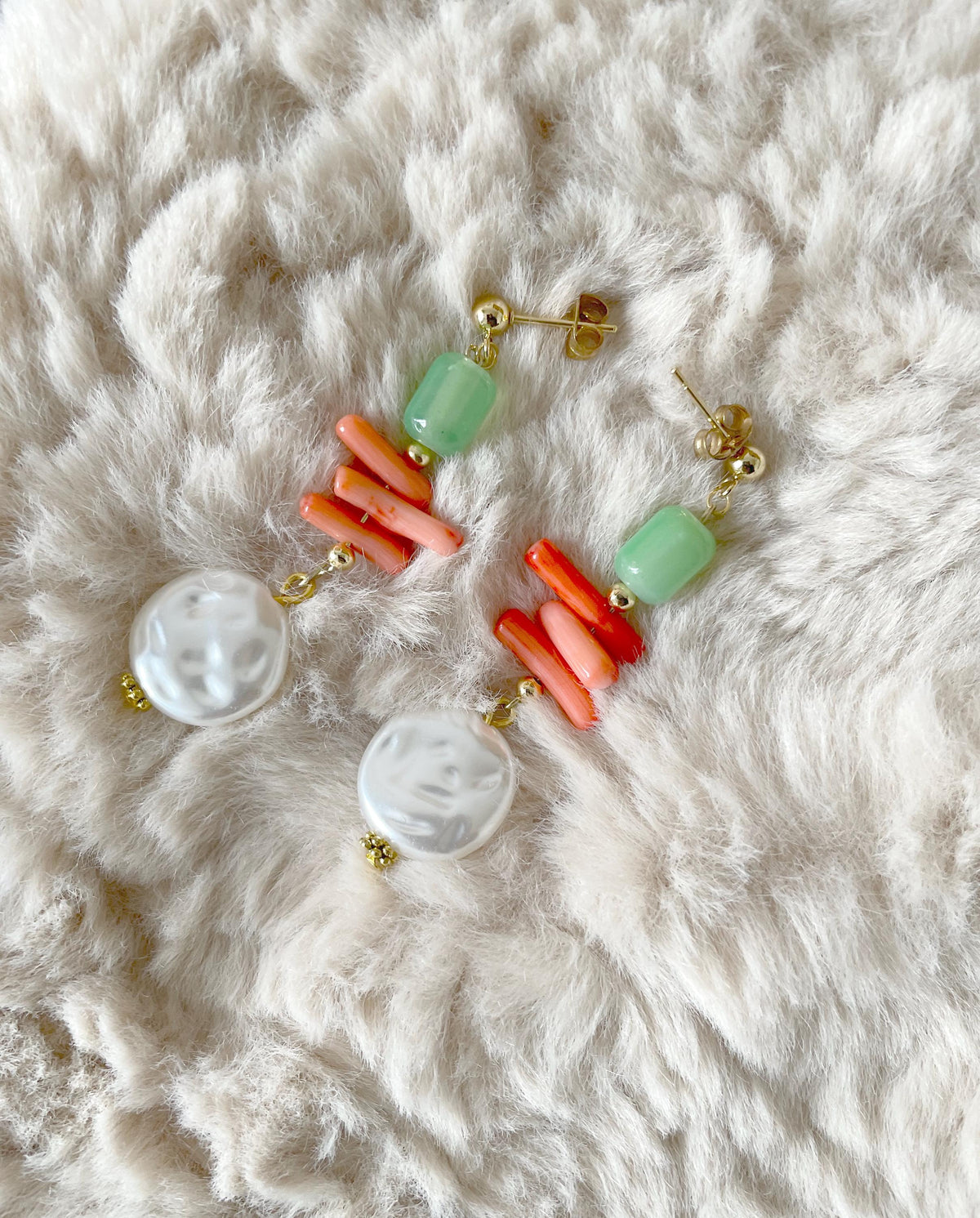 A pair of bohemian-style drop earrings featuring light green beads, orange and coral tube beads, white baroque pearls, and gold-tone posts, displayed on a textured white faux fur background.