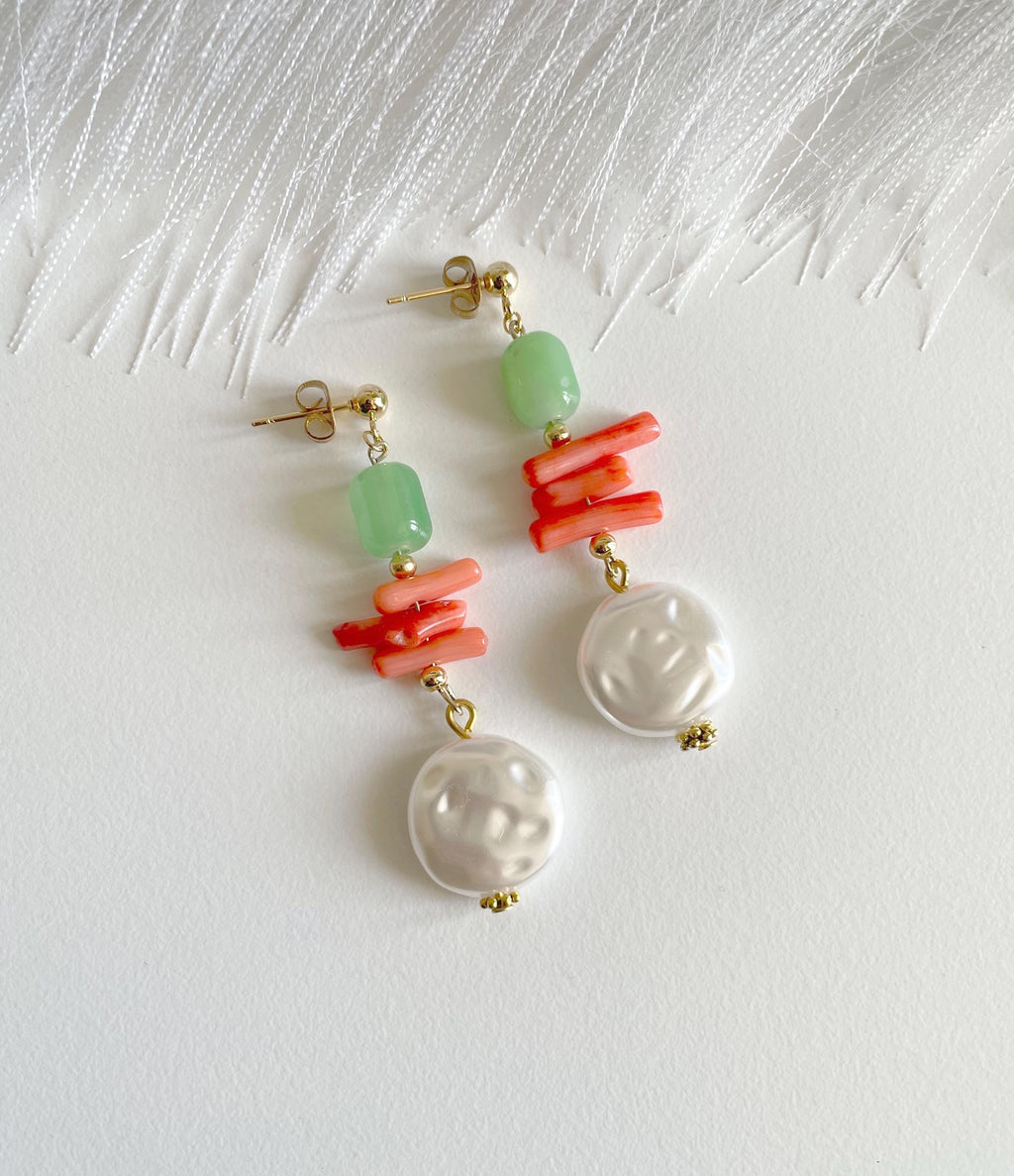 A pair of drop earrings with gold-tone posts, light green beads, coral-red stacked bars, and white baroque pearl dangles, displayed on a textured white surface.