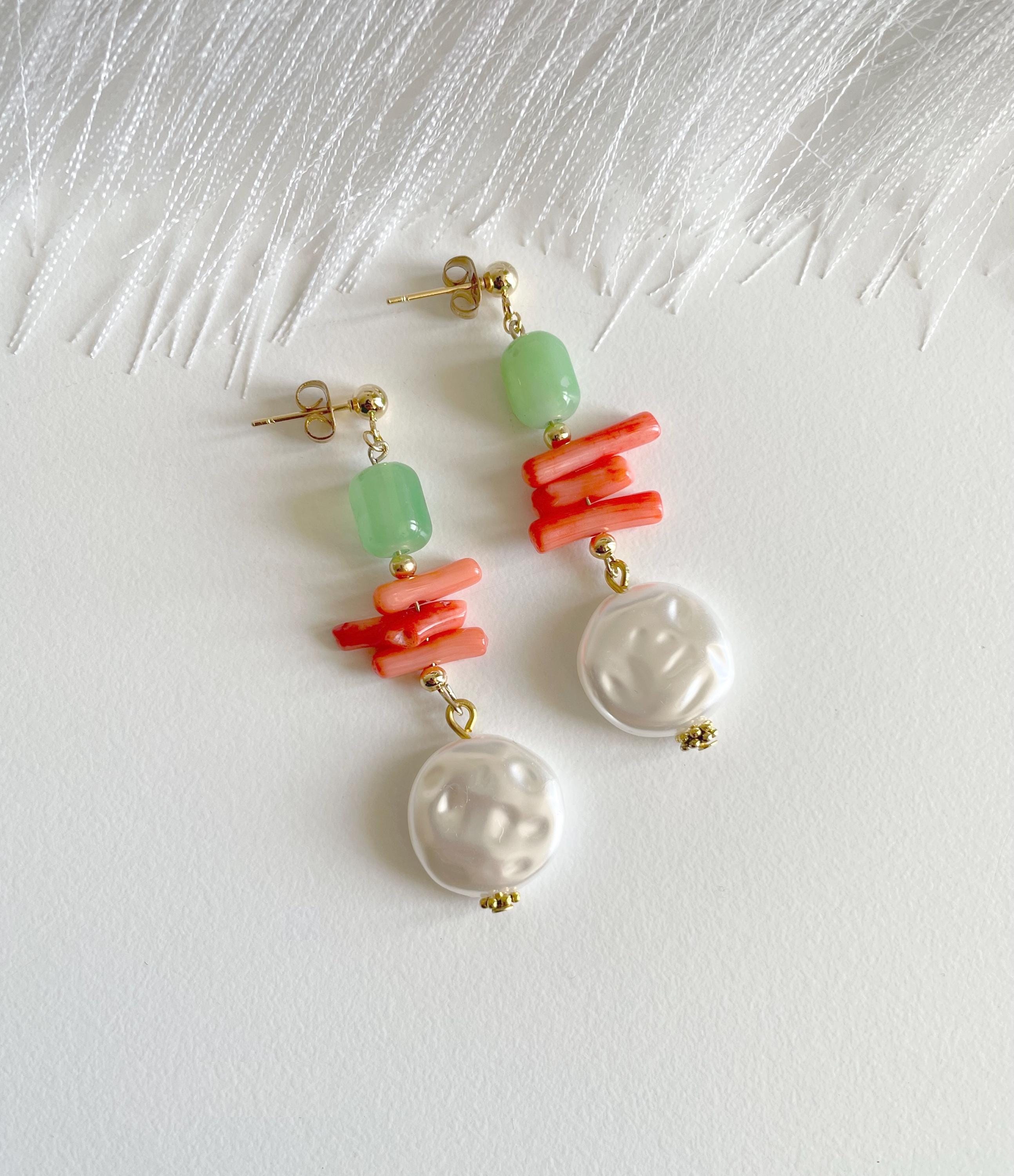 A pair of drop earrings with gold-tone posts, light green beads, coral-red stacked bars, and white baroque pearl dangles, displayed on a textured white surface.