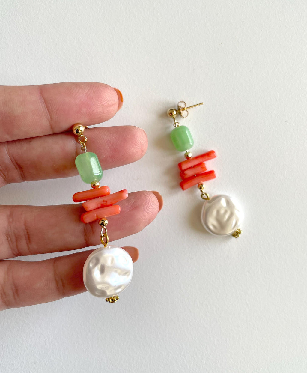 A pair of drop earrings held in a person's hand against a plain off-white background. Each earring has a gold post, a light green faceted bead, a cluster of bright coral-orange cylindrical beads, a white baroque pearl, and a larger irregular white pearl at the bottom.
