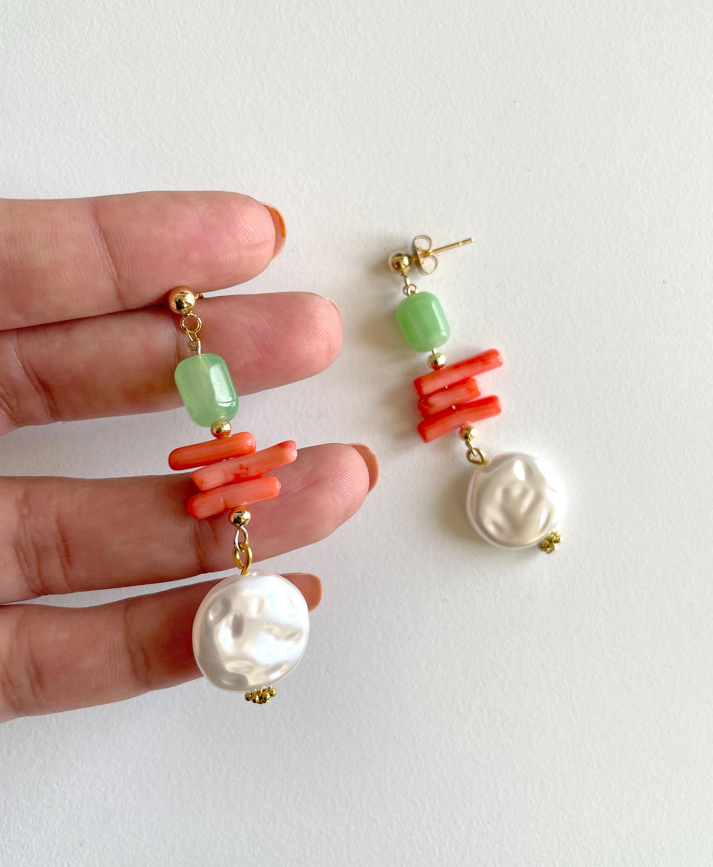 A pair of drop earrings held in a person's hand against a plain off-white background. Each earring has a gold post, a light green faceted bead, a cluster of bright coral-orange cylindrical beads, a white baroque pearl, and a larger irregular white pearl at the bottom.