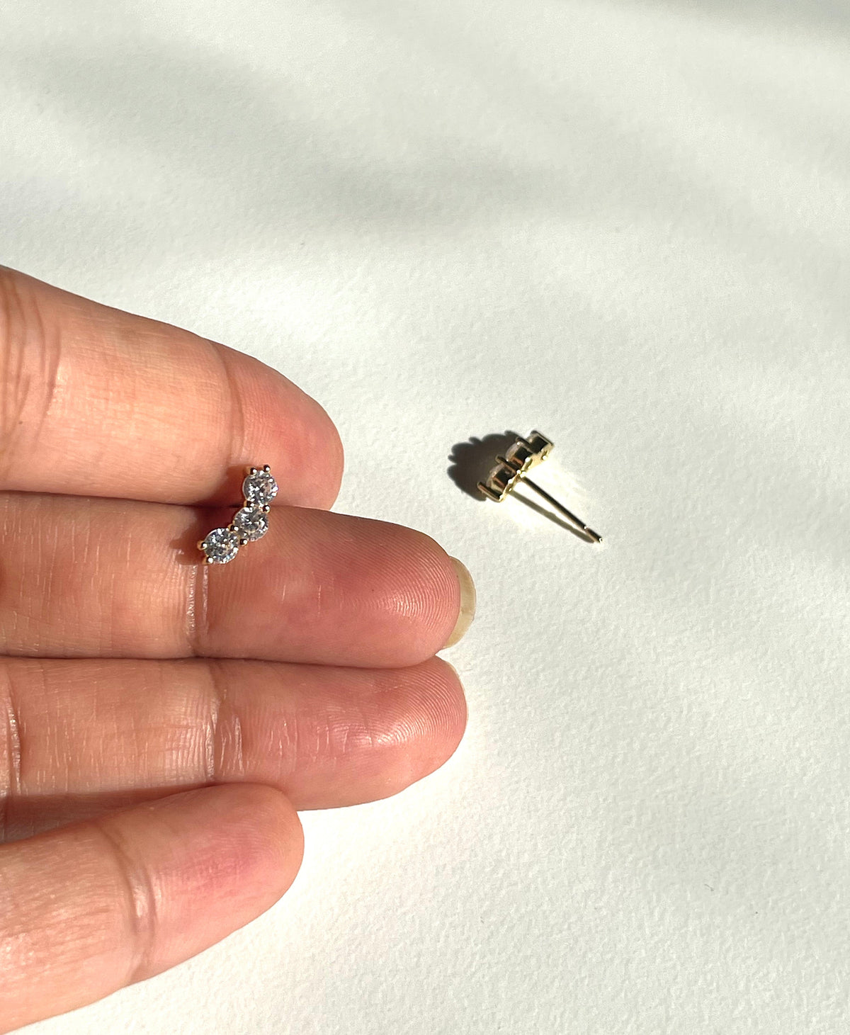 A close-up of a hand holding a small gold plated curved stud earring with clear stones, on a white textured background.