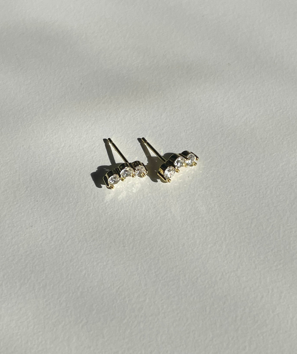 A pair of gold plated stud earrings with three clear round-cut gemstones each, displayed on a textured off-white surface with soft shadows.