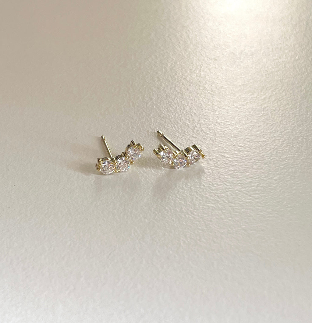 A pair of gold stud earrings with clear round-cut gemstones, displayed on a textured off-white surface.