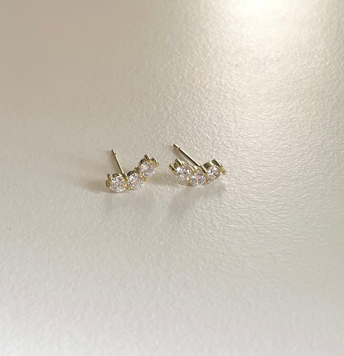 A pair of gold stud earrings with clear round-cut gemstones, displayed on a textured off-white surface.