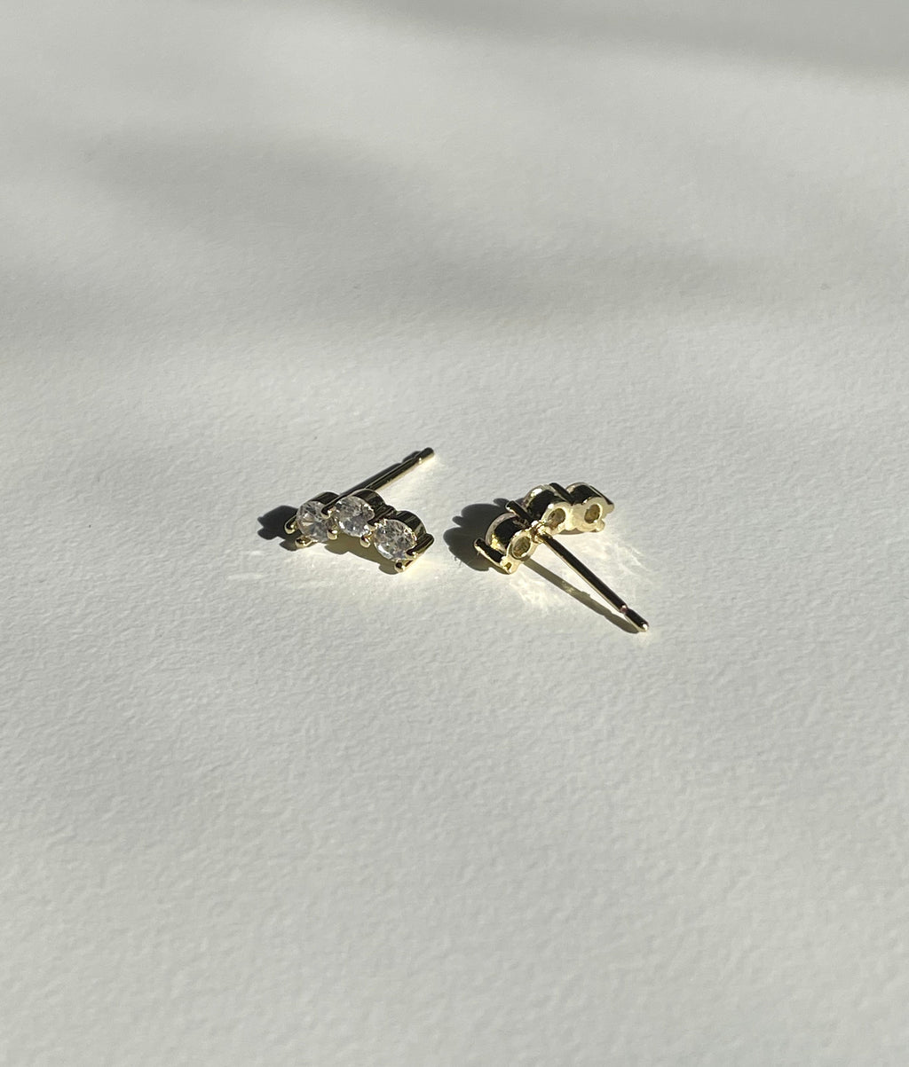 A pair of small gold plated stud earrings with clear gemstones, shown from the back on a textured white surface with soft shadows.