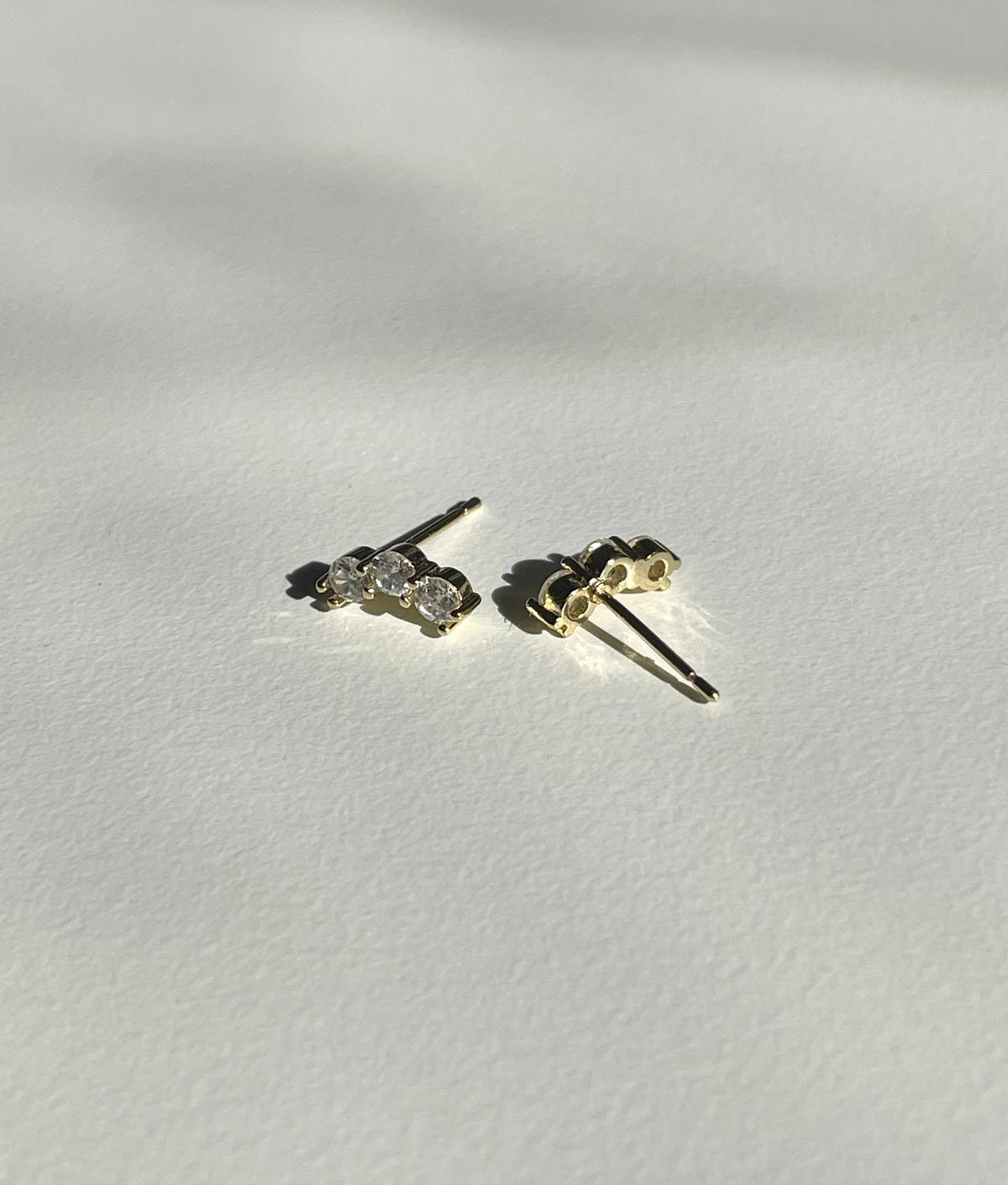 A pair of small gold plated stud earrings with clear gemstones, shown from the back on a textured white surface with soft shadows.
